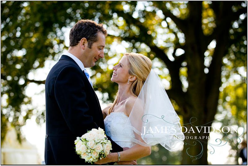 Bride in white dress and groom in suit outdoors, surrounded by trees and sunlight.