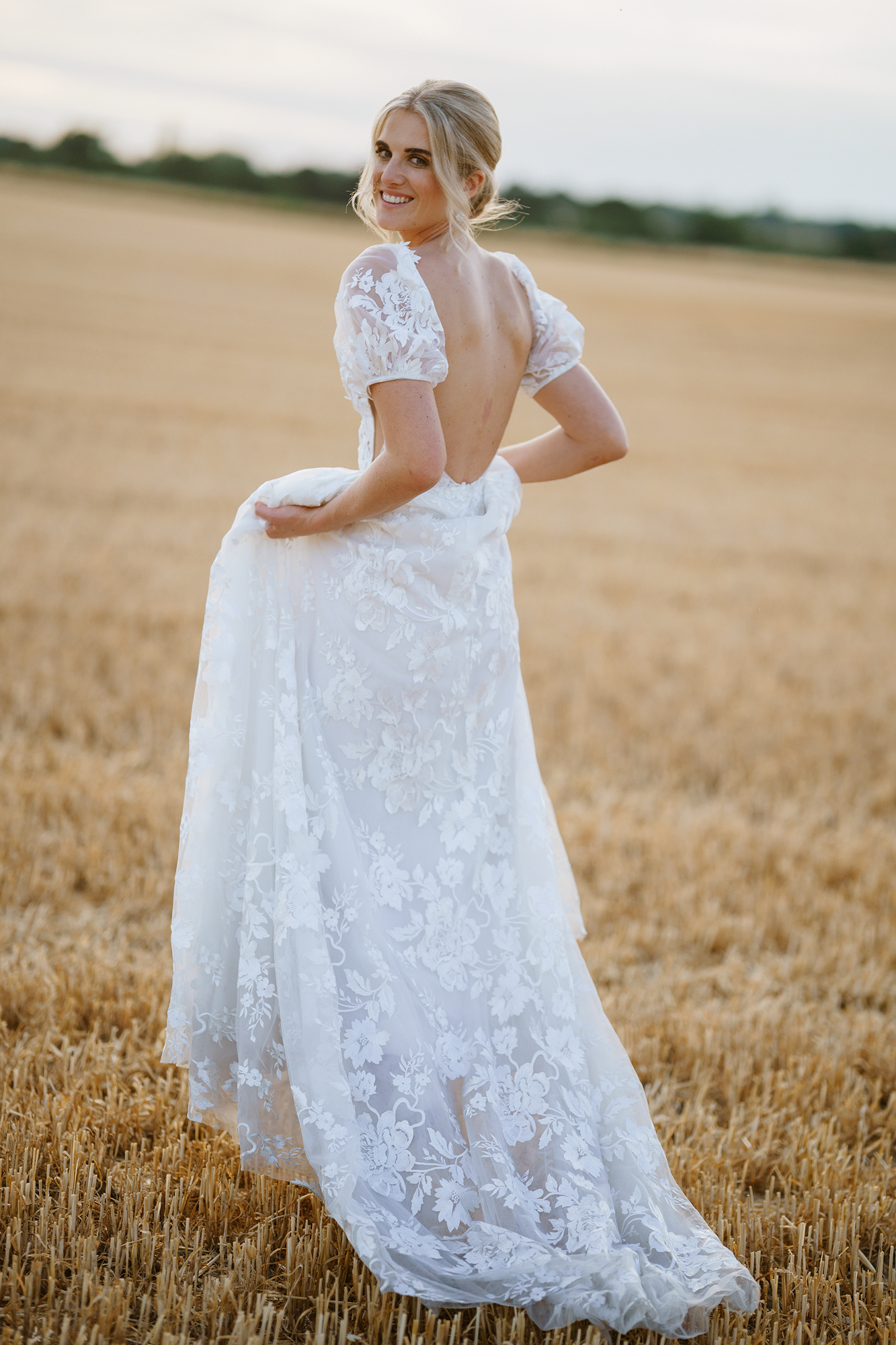 Person in backless white lace dress stands in harvested field with distant tree line.