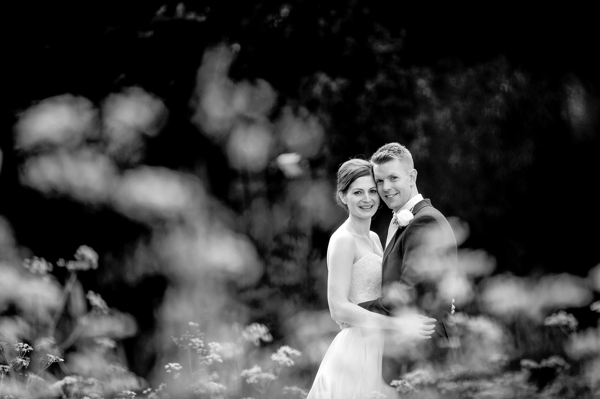 Couple in wedding attire posing outdoors with blurred foliage, captured in black and white.