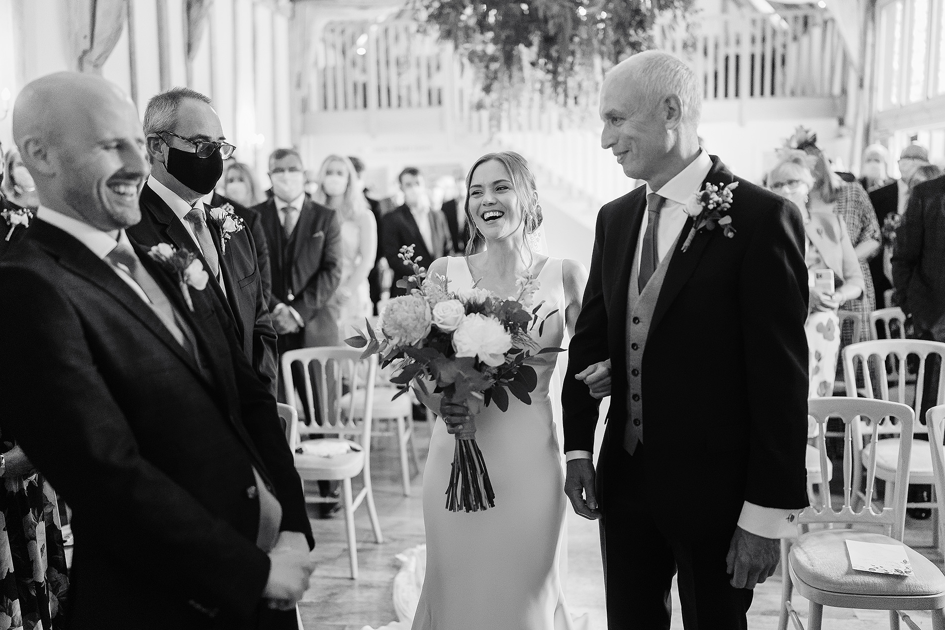 Bride with bouquet smiling between two men in suits at a wedding ceremony with seated guests.