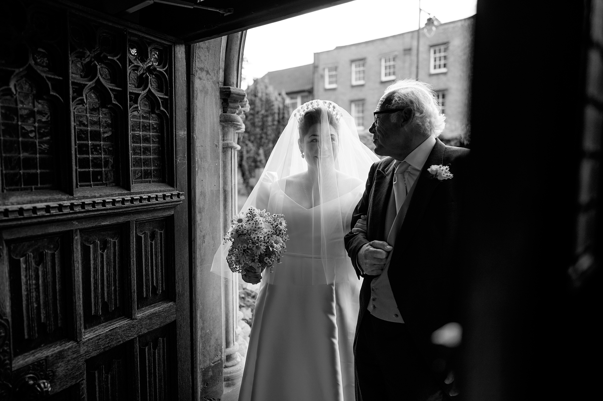 Bride with bouquet and older man in doorway, black and white, building in background.