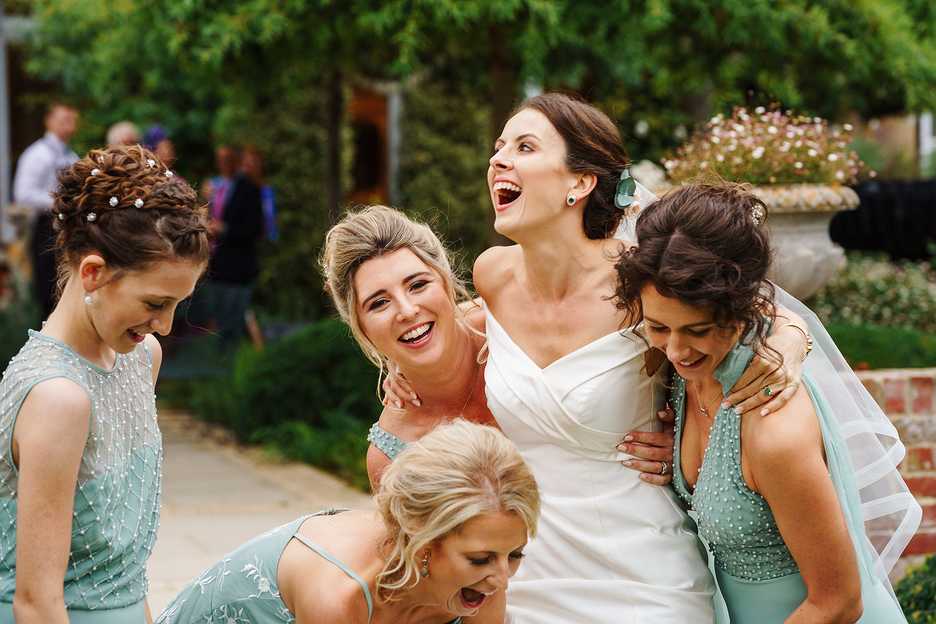 Women in formal dresses, including a bride, joyfully celebrating outdoors with greenery around.