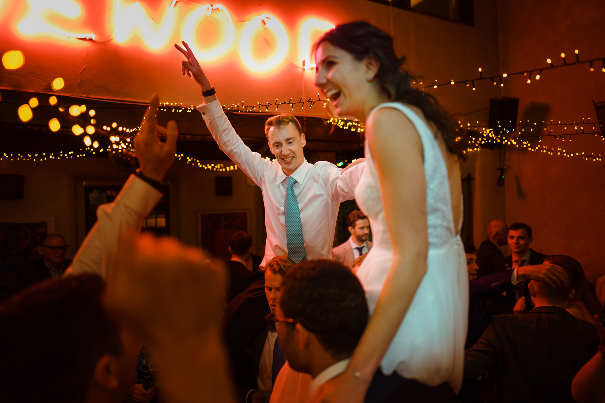 Bride and groom lifted on shoulders at festive indoor celebration with string lights.