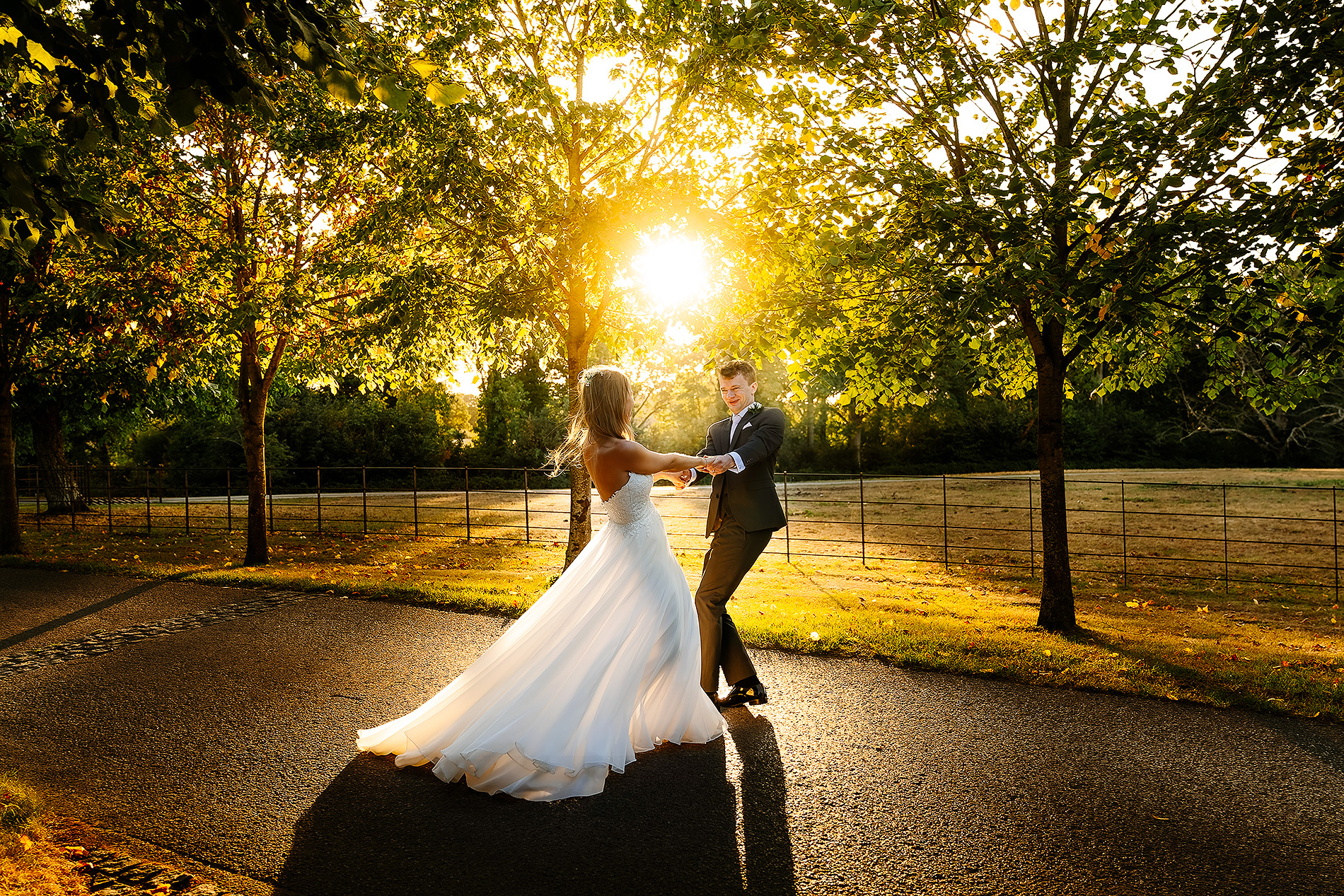 Bride and groom dance on sunlit path, surrounded by trees with a warm sunset glow.