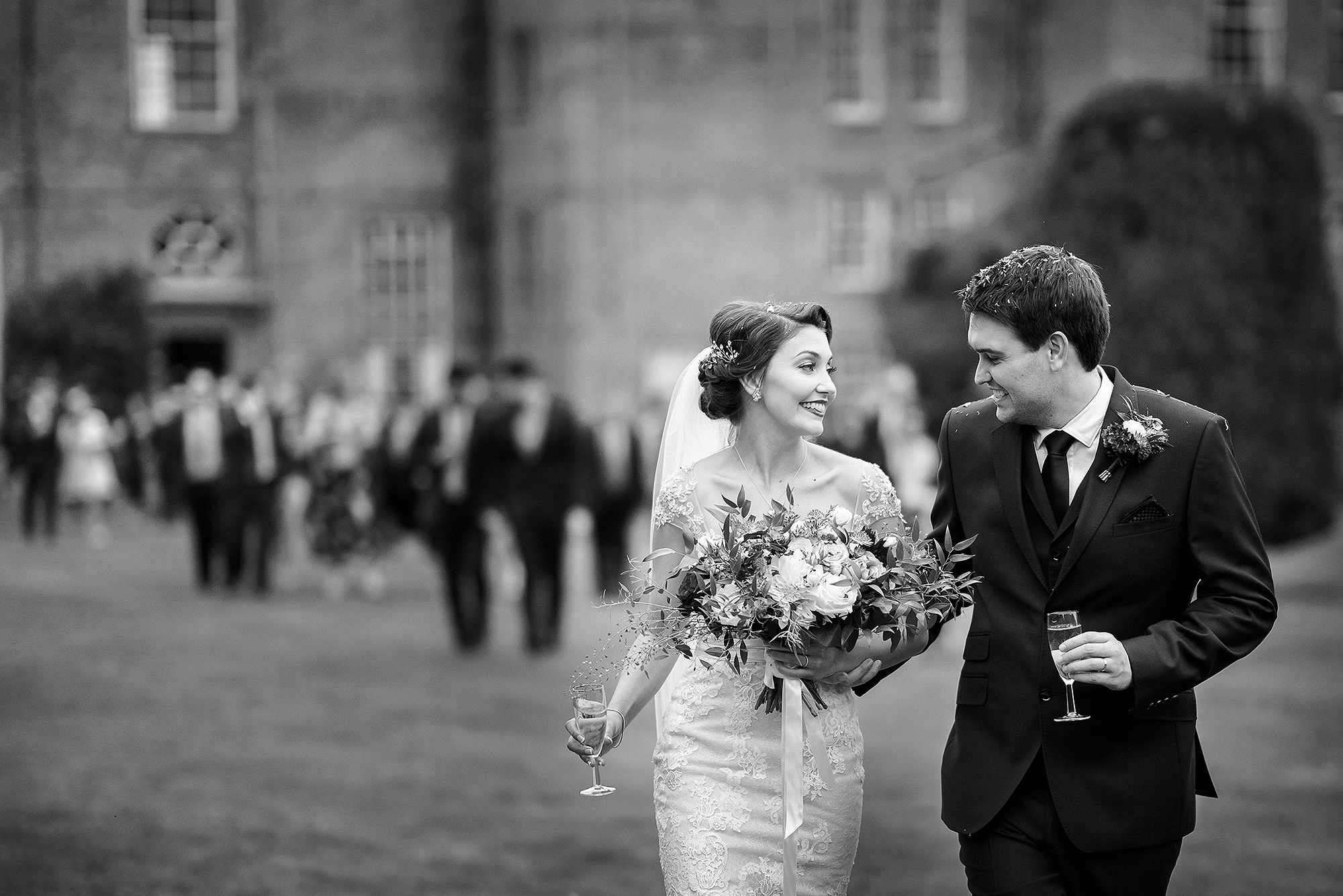 Bride and groom smiling, holding bouquet and glasses, walk with guests in garden setting.