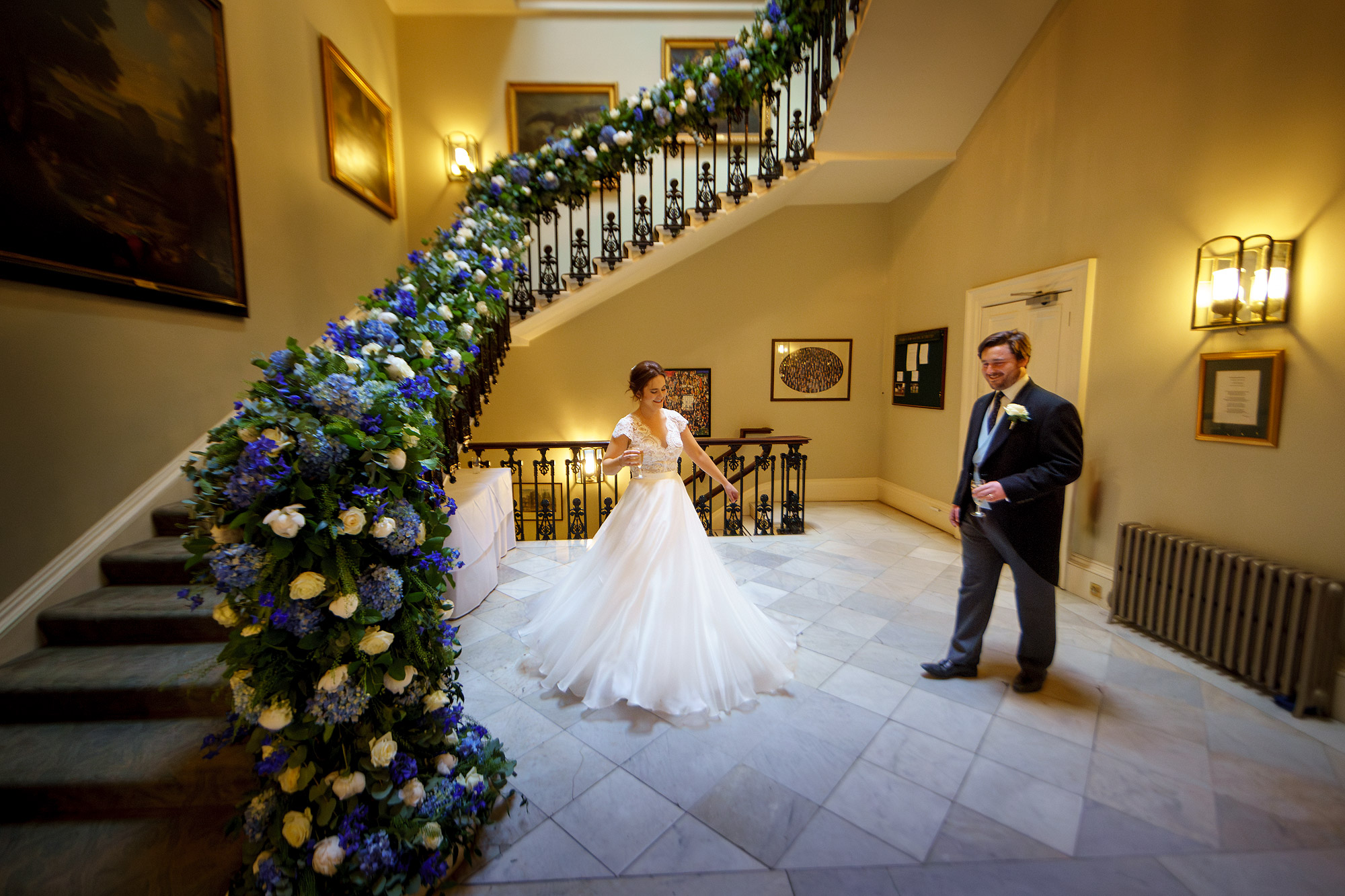 Bride and groom by grand staircase with blue and white flowers in elegant interior.