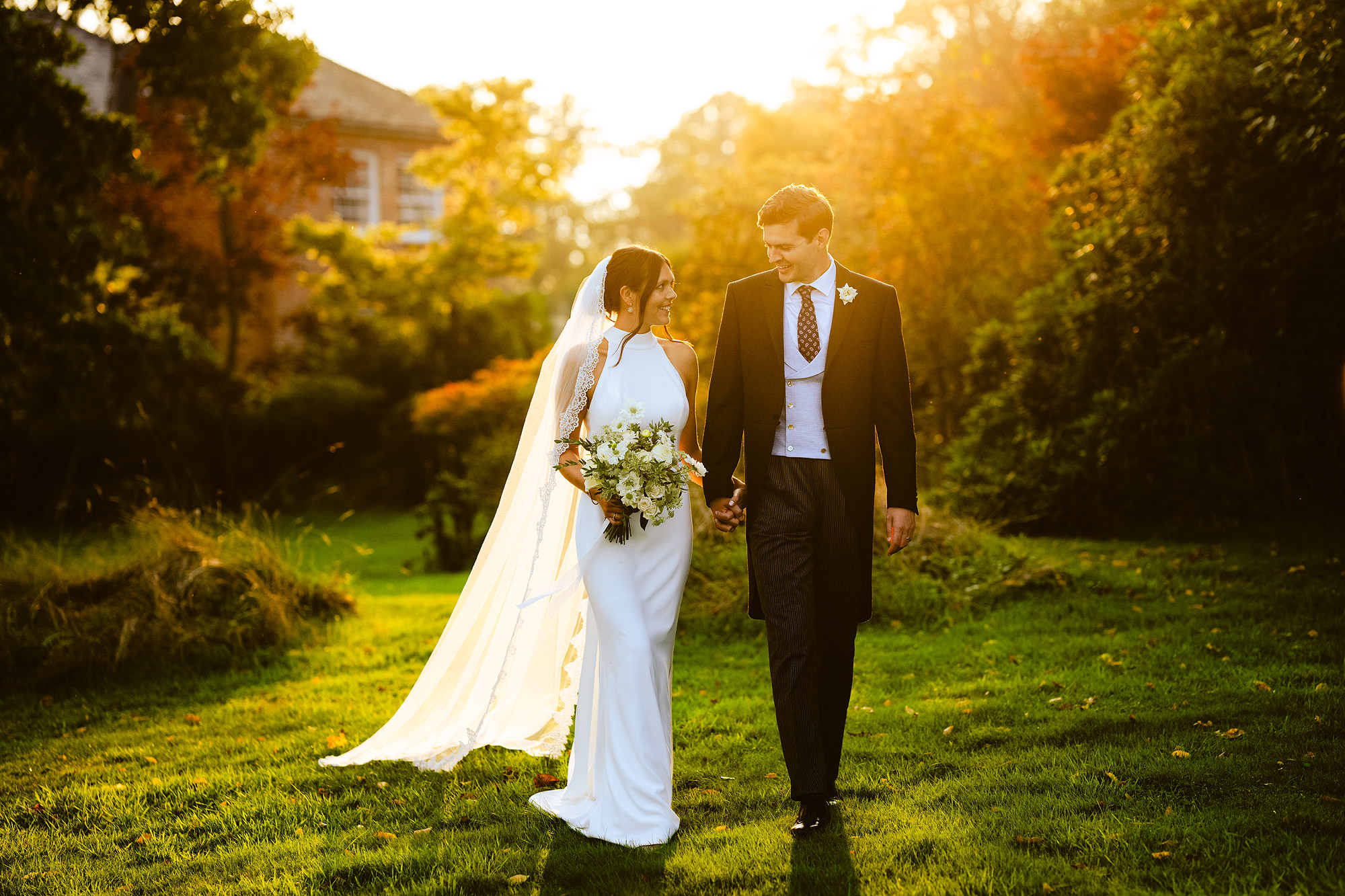 Bride and groom walk hand in hand on a lawn, with trees and sunlight around them.