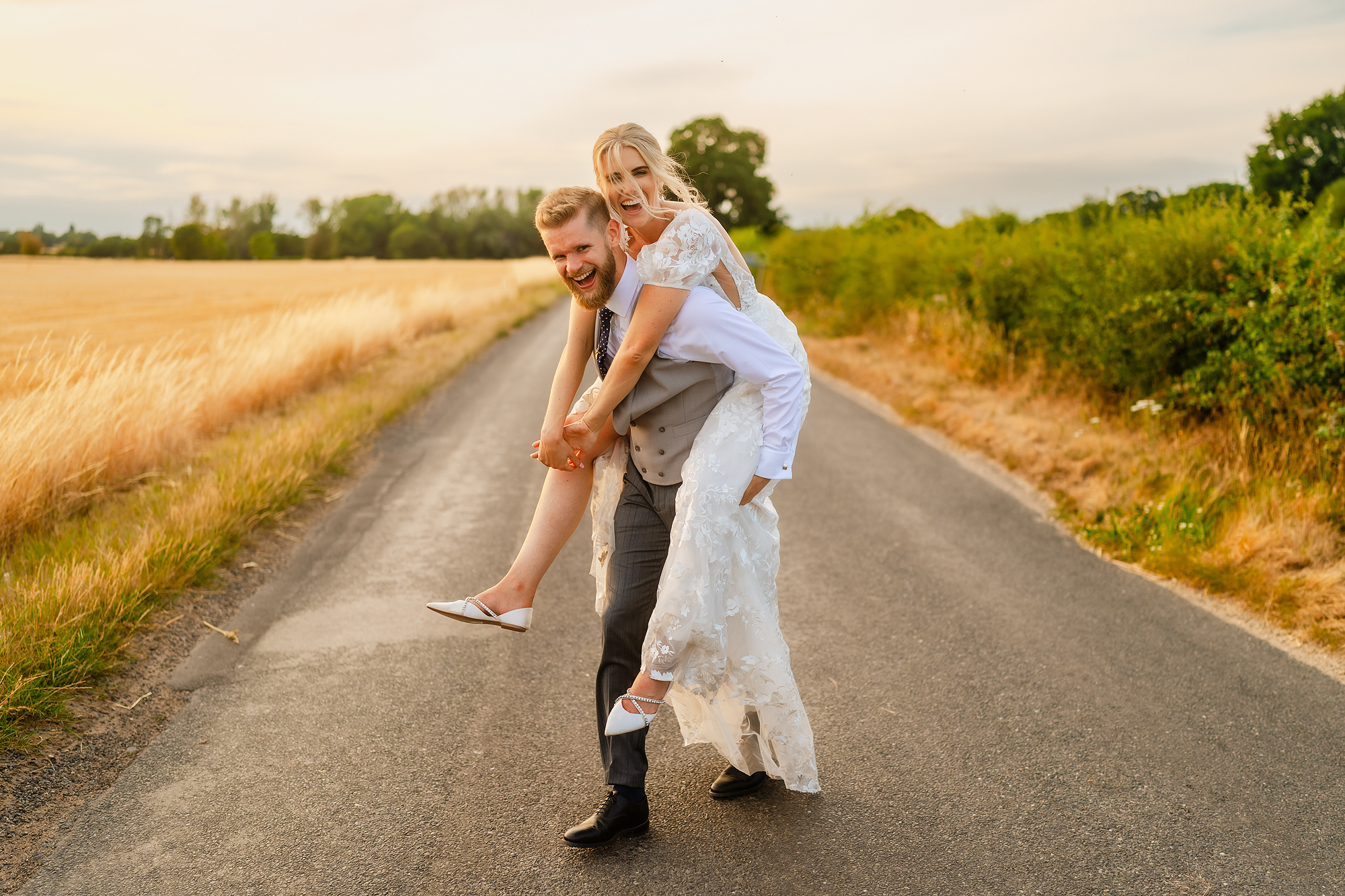 Bride in white dress carried by groom on rural road, fields and clear sky background.