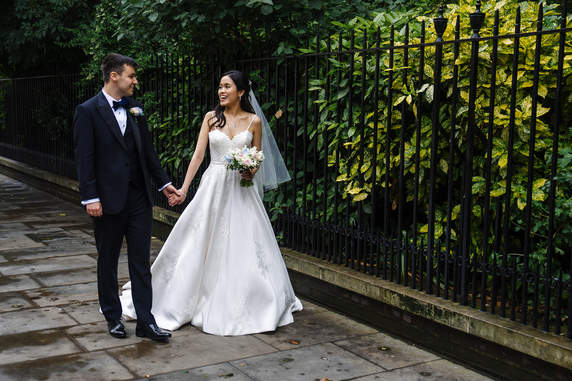 Couple walking hand in hand by a fence, bride in white dress with bouquet, groom in dark suit.
