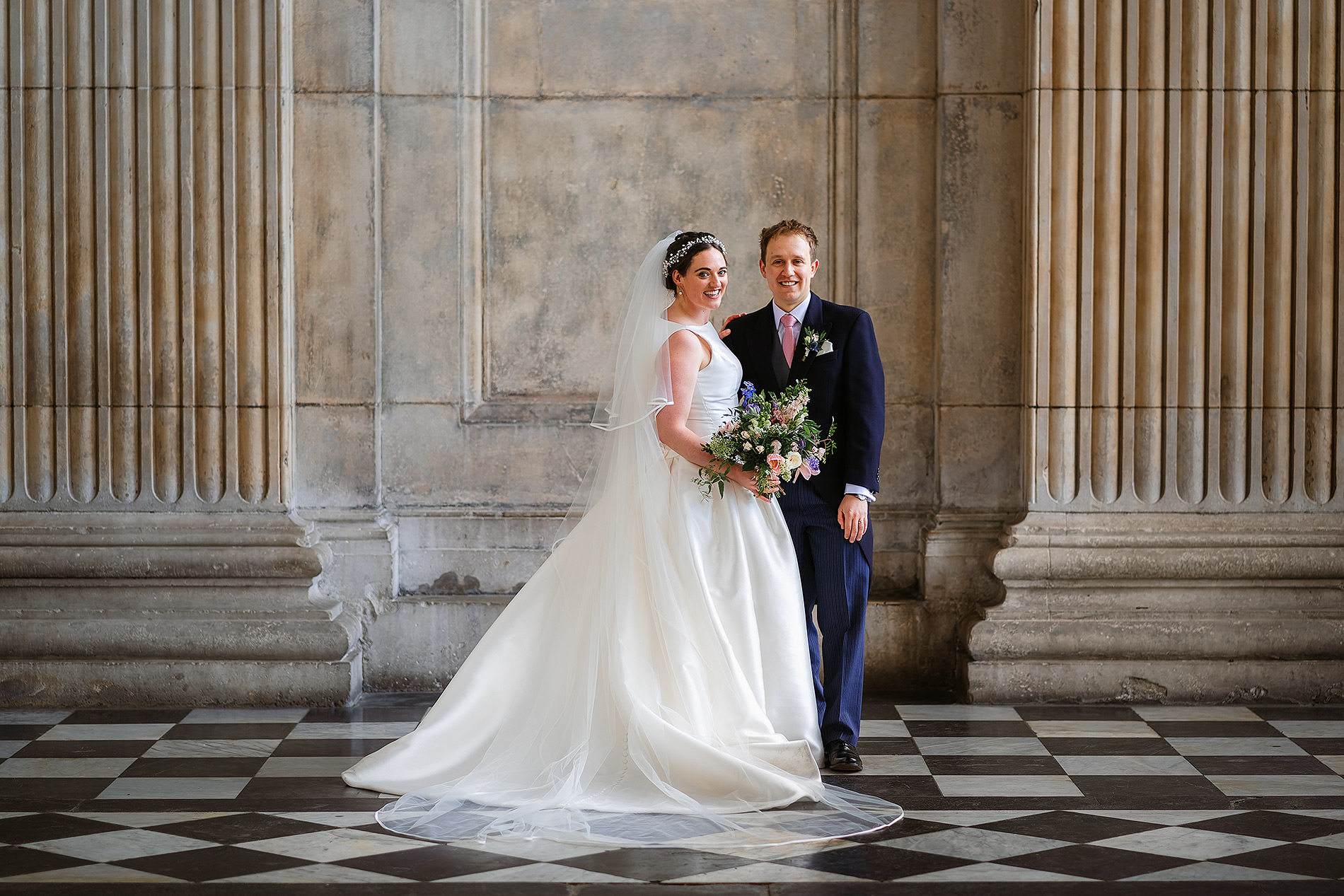 Bride and groom smile in wedding attire with bouquet, between large stone columns on tiled floor.