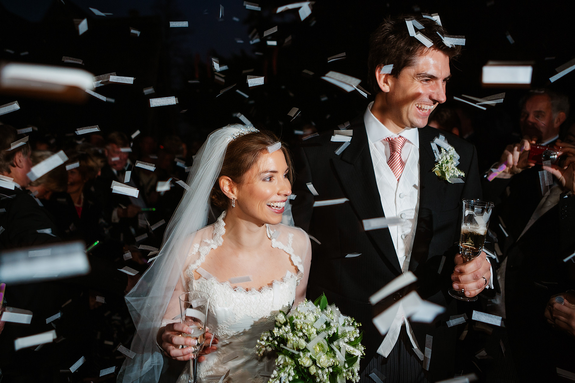 Bride and groom smiling under confetti, holding glasses and bouquet at wedding celebration.