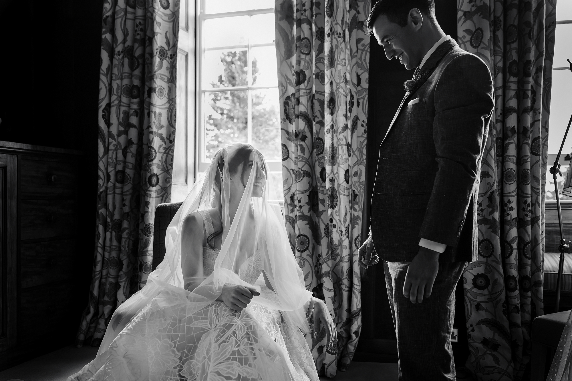 Bride seated in gown and veil, groom stands nearby; room with patterned curtains and window.