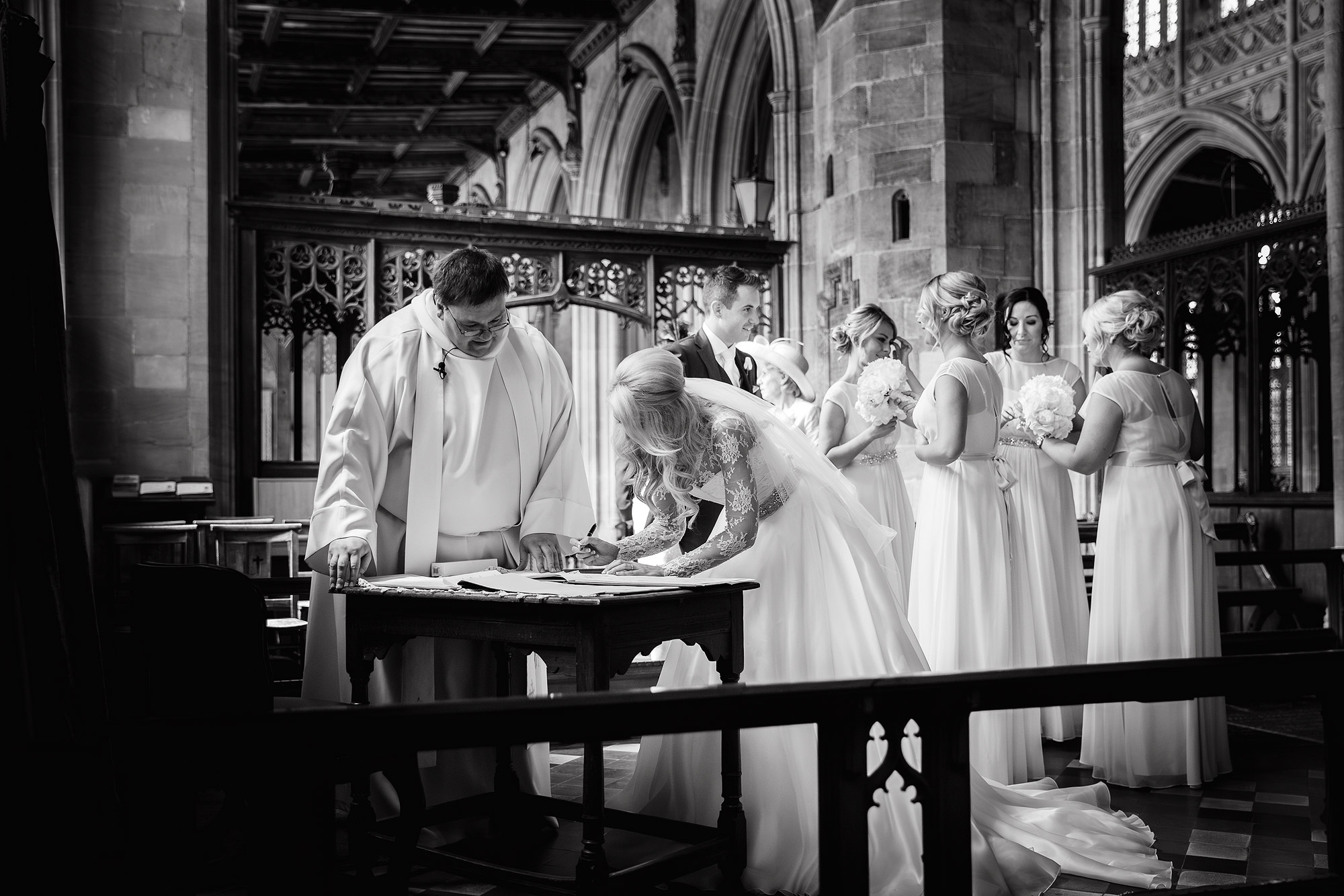 Bride signing document in a church with clergy, bridesmaids, groom, and ornate architecture.