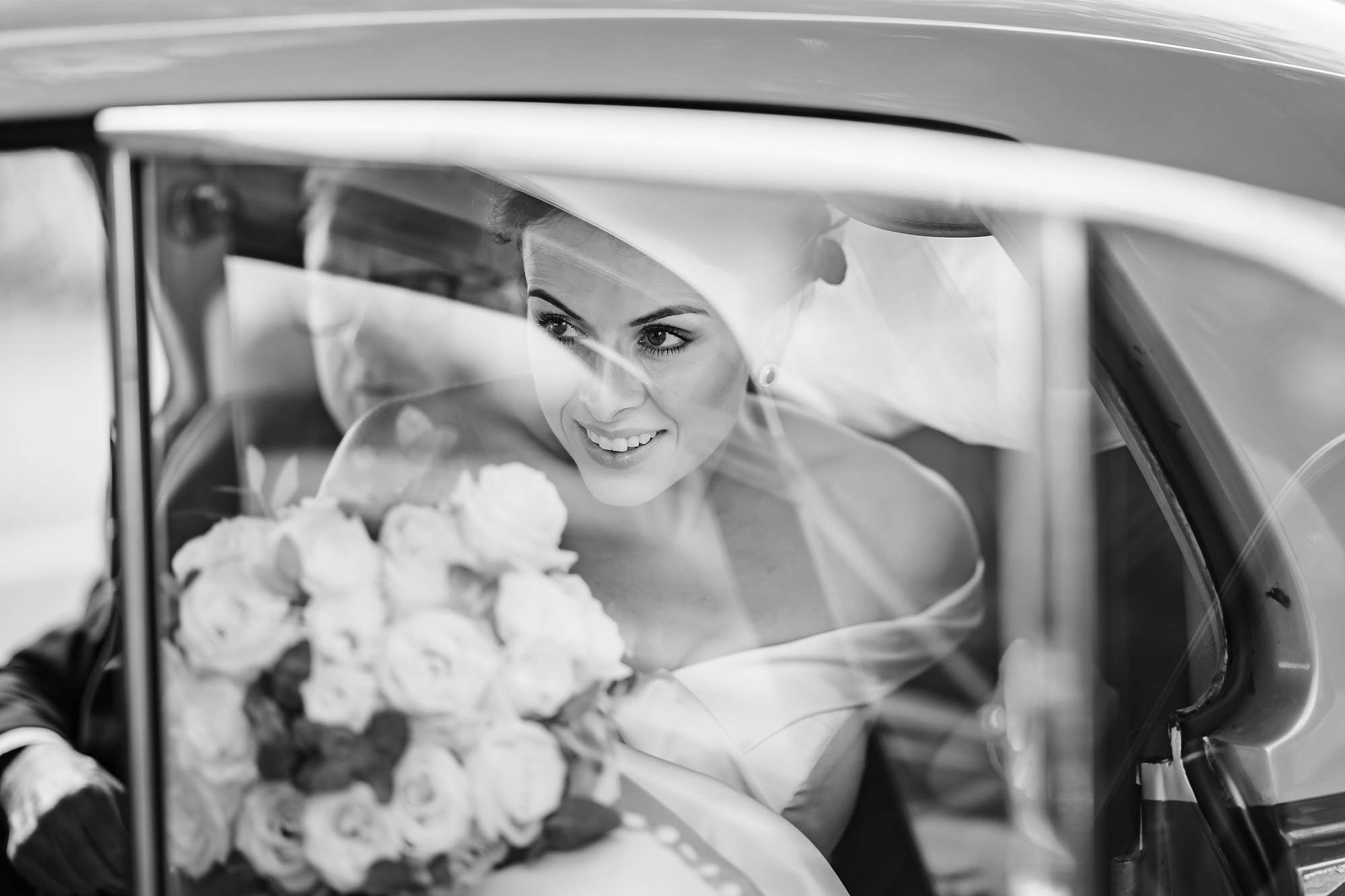 Bride in wedding dress with bouquet inside a car, seen through window in black and white photo.
