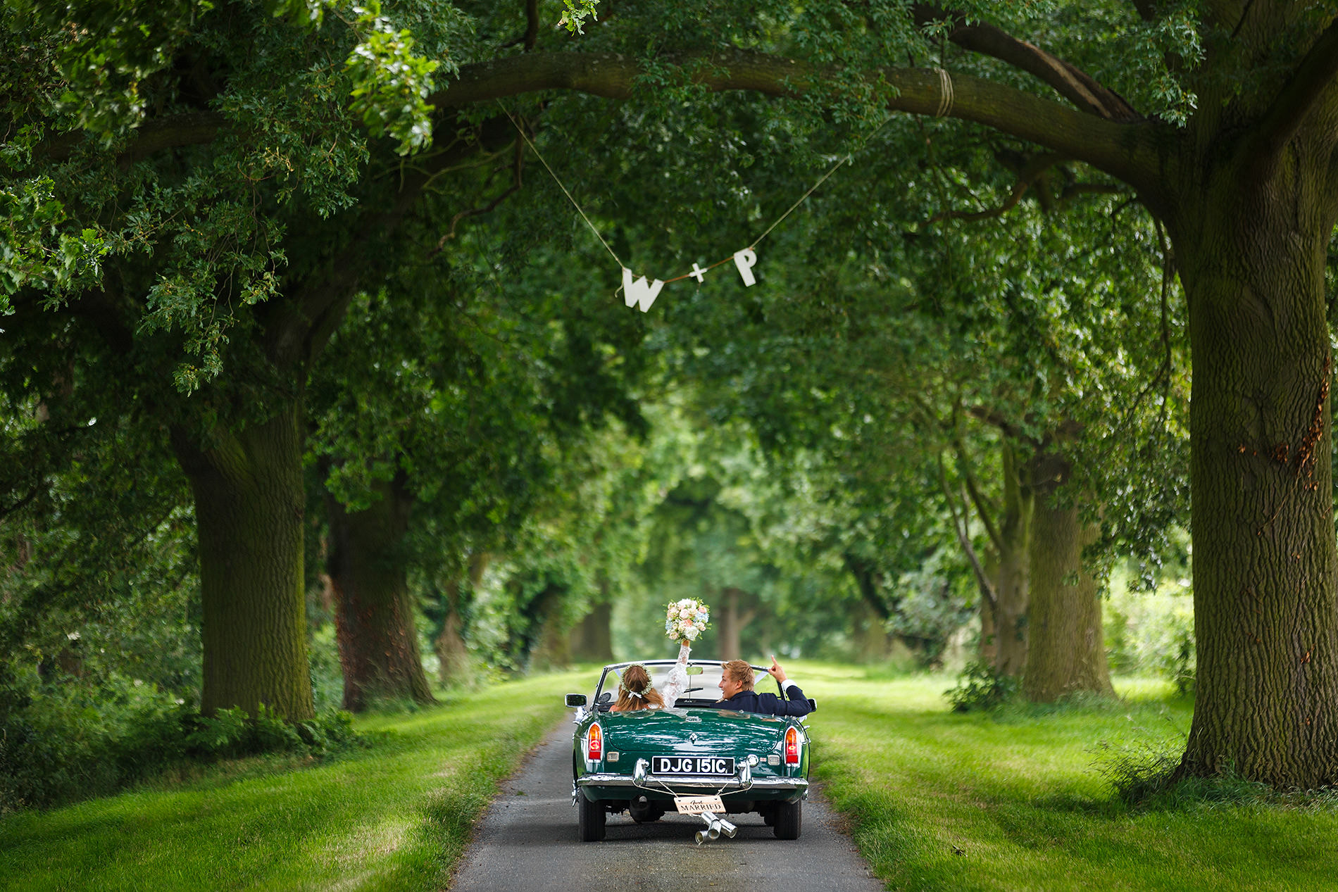 Classic convertible on tree-lined road, two people inside, one waving flowers, "W + P" banner above.