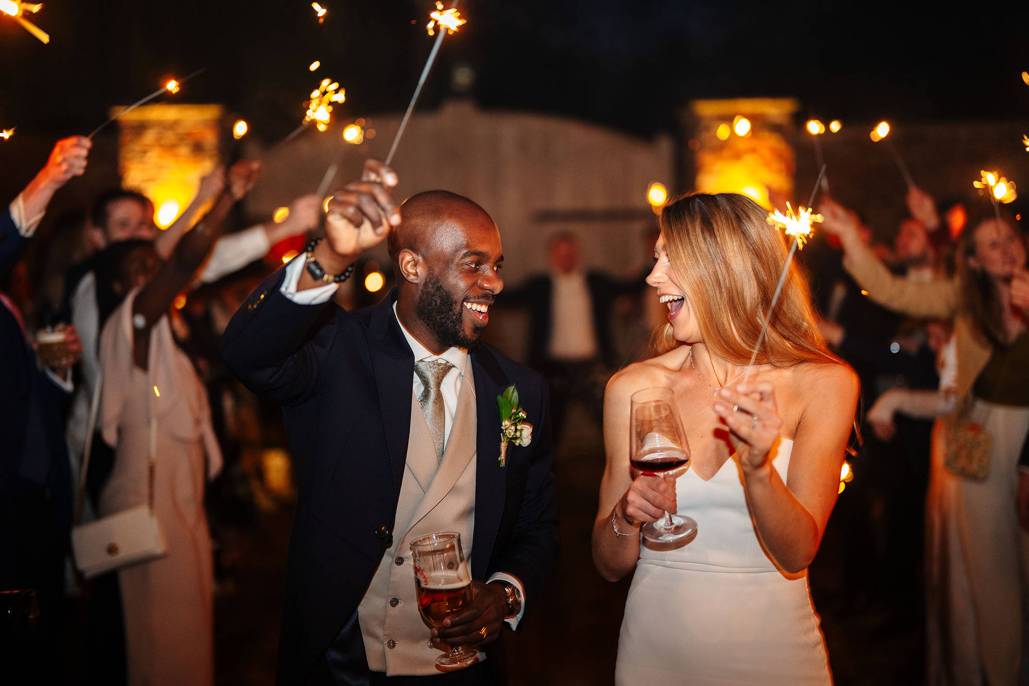 Couple in formal attire celebrating outdoors with sparklers and drinks, surrounded by guests.