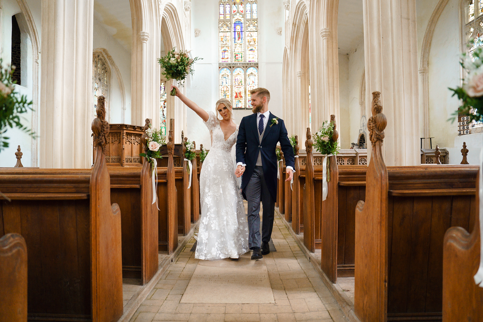 Couple walking down a church aisle in wedding attire, bride lifting bouquet, stained glass behind.