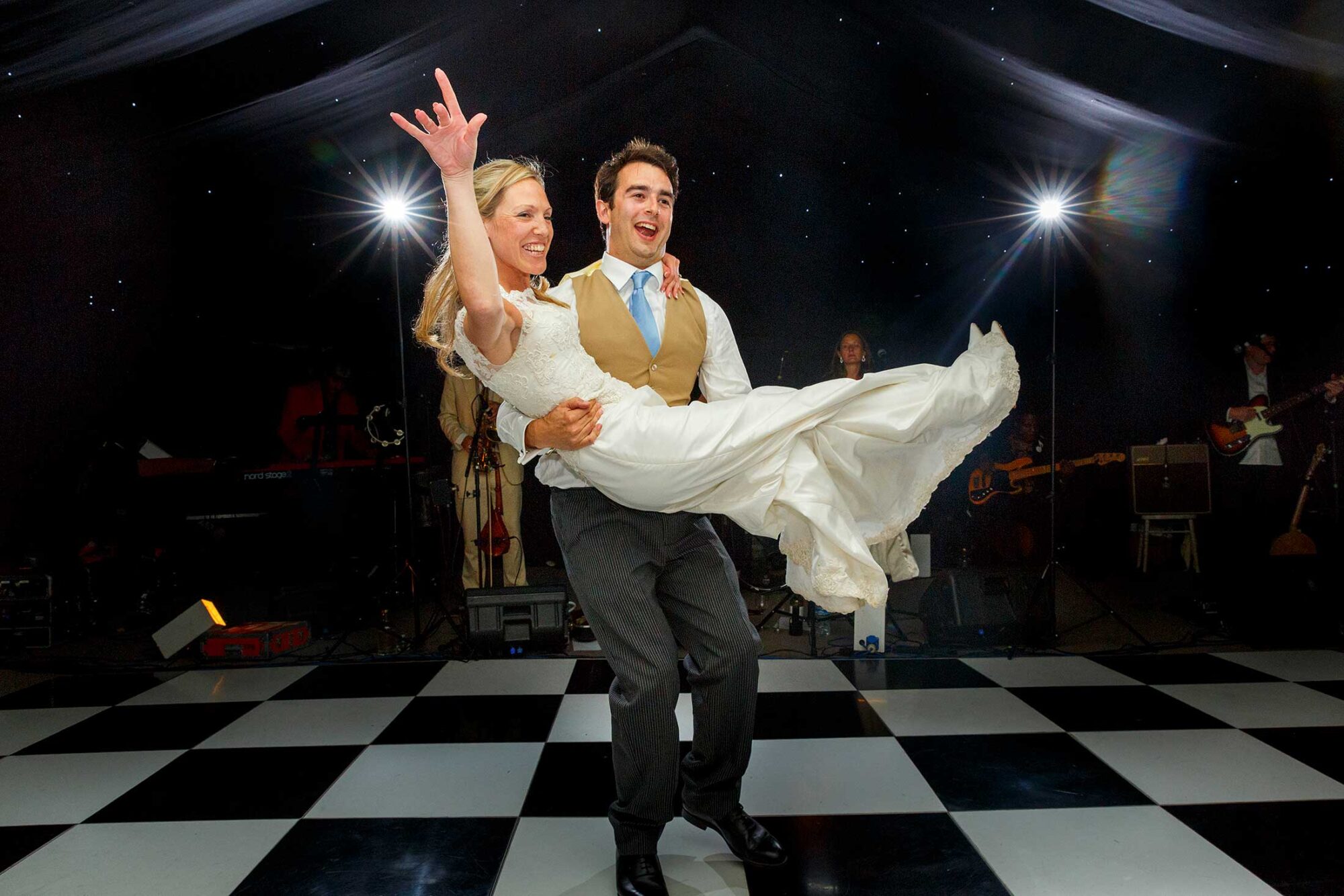 Couple dancing on checkered floor; man lifts woman in white dress; musicians in background.