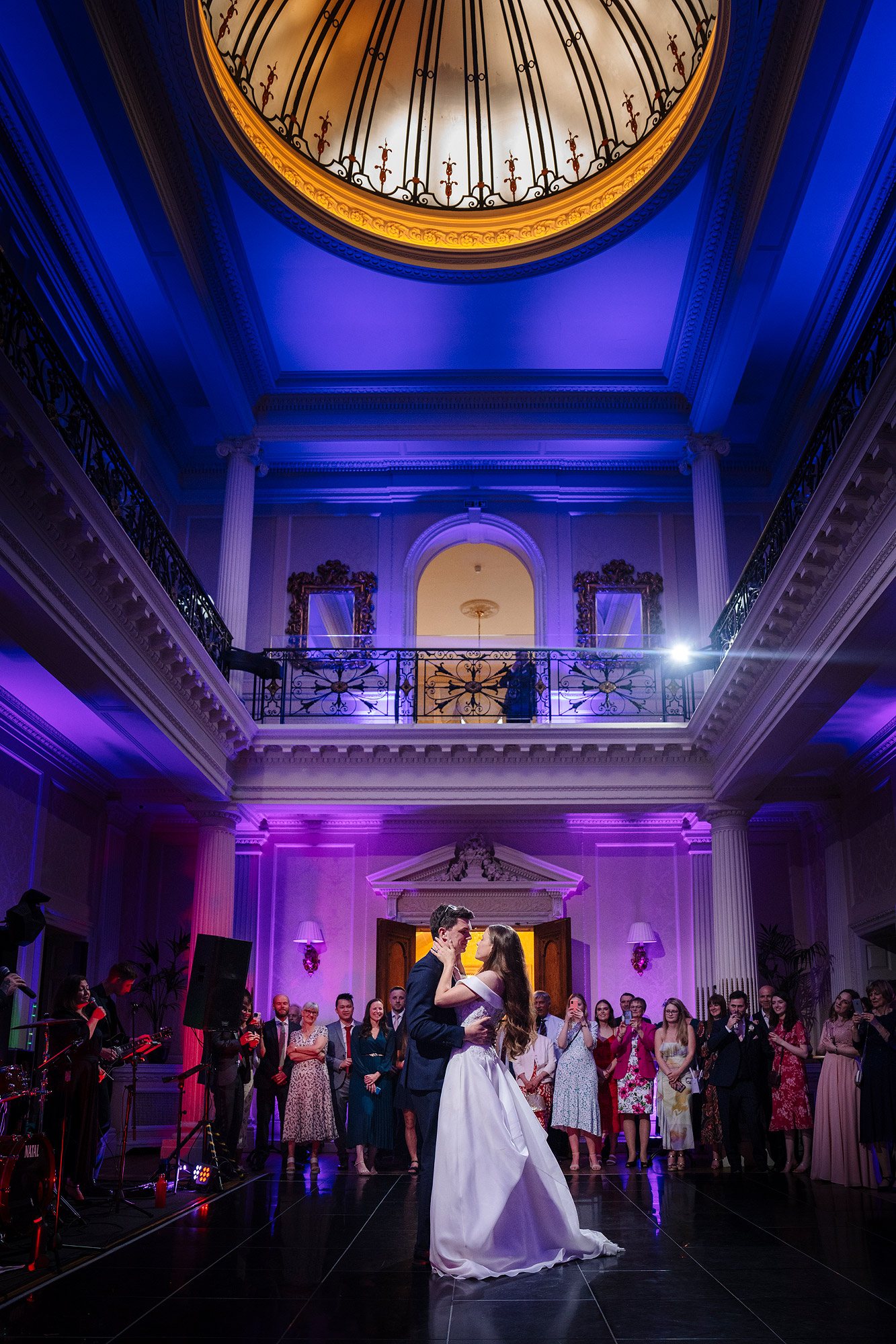 Couple dancing in an elegant room with a large crowd of guests watching under a circular skylight.