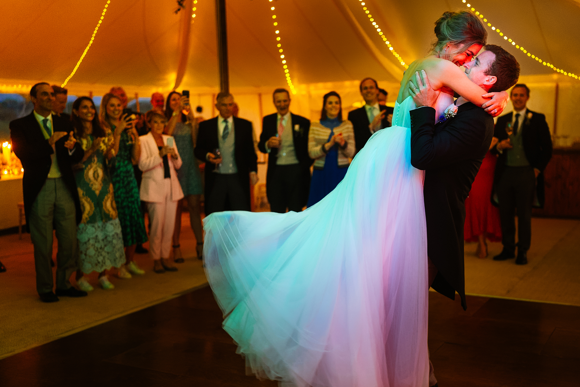 Couple dancing at wedding under tent with string lights, surrounded by guests taking photos.