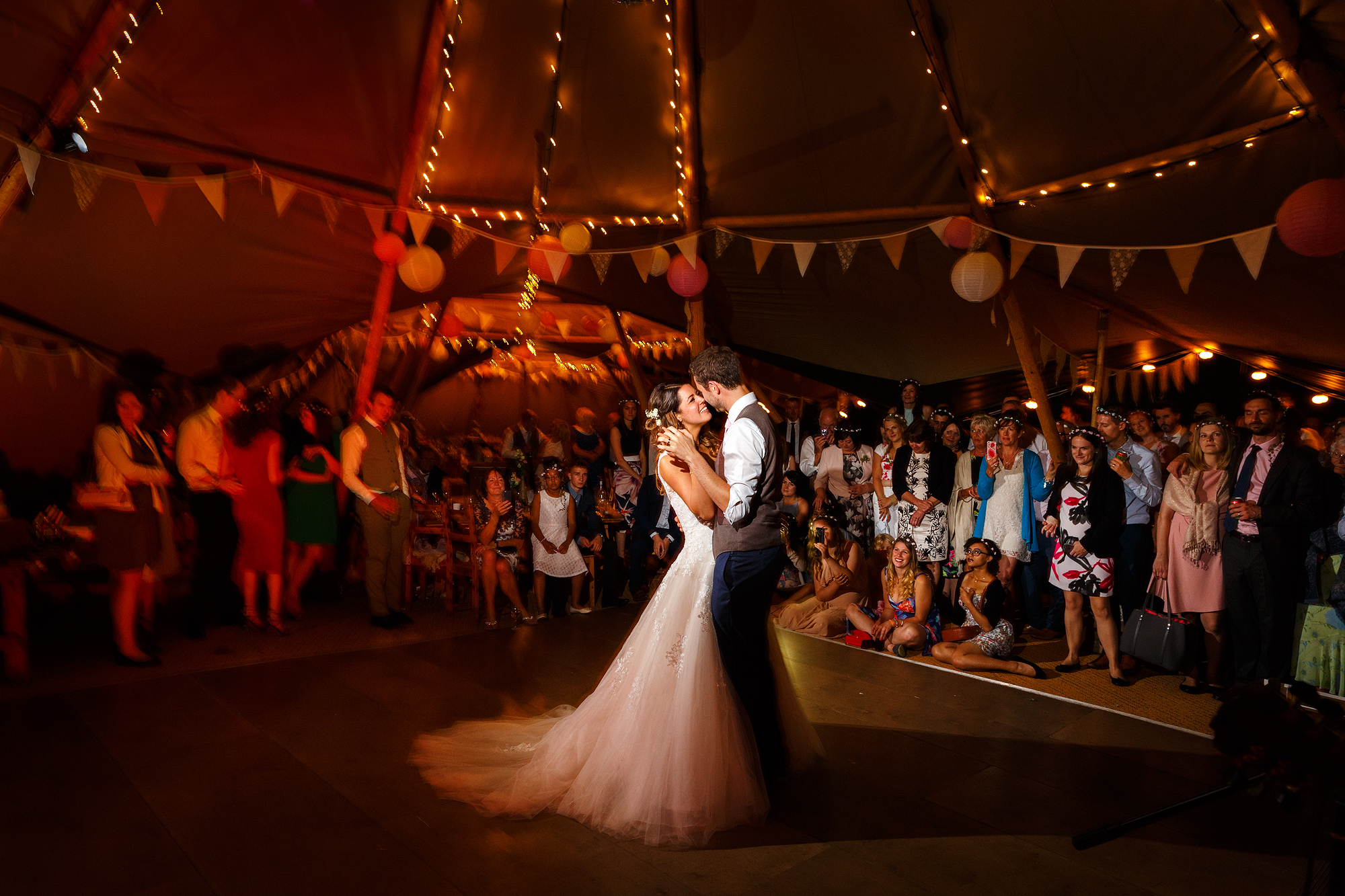 Couple dances at wedding in decorated tent with string lights, bunting, and guests watching.