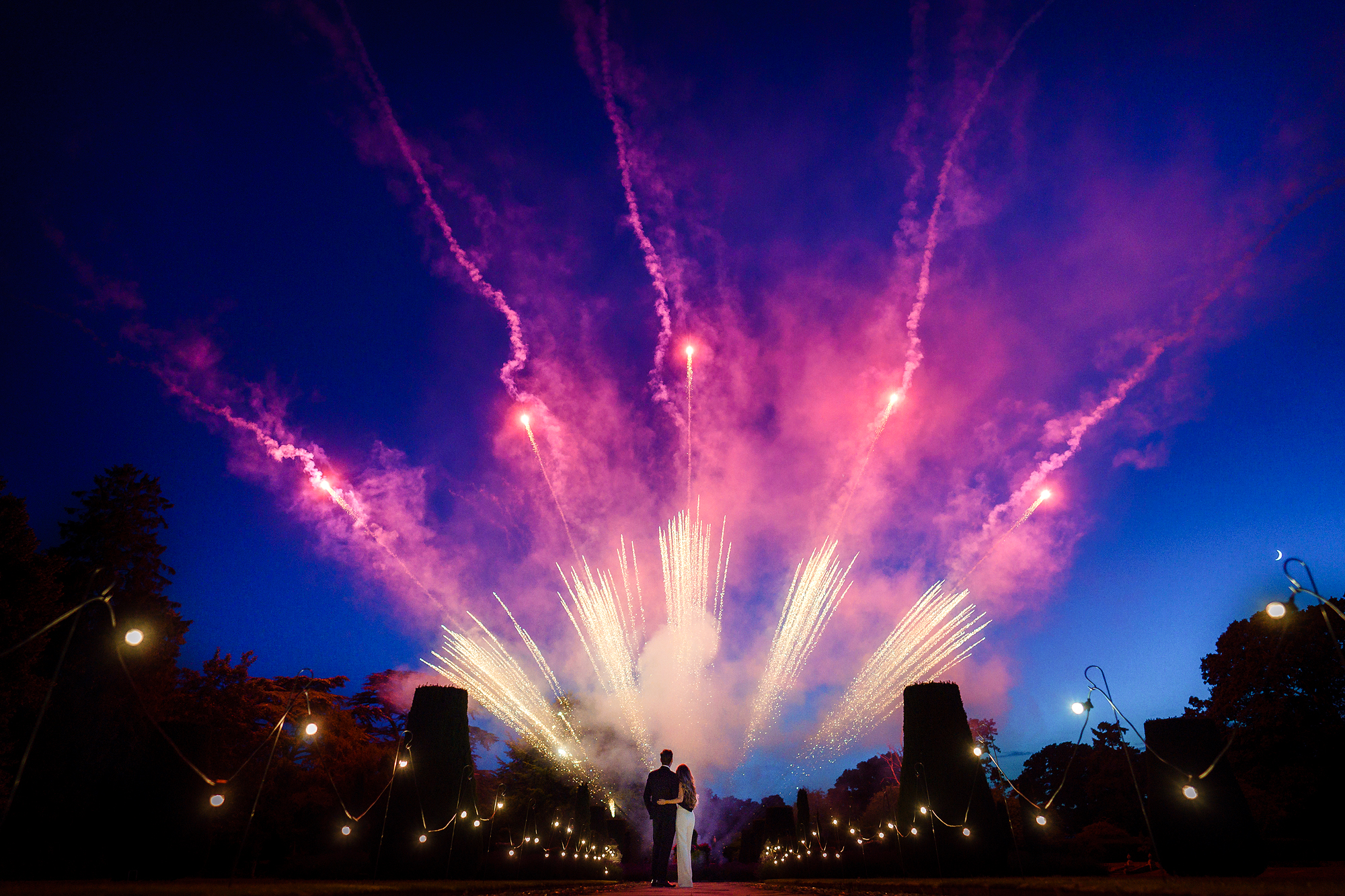 Couple embracing outdoors at night with fireworks and string lights in the sky.