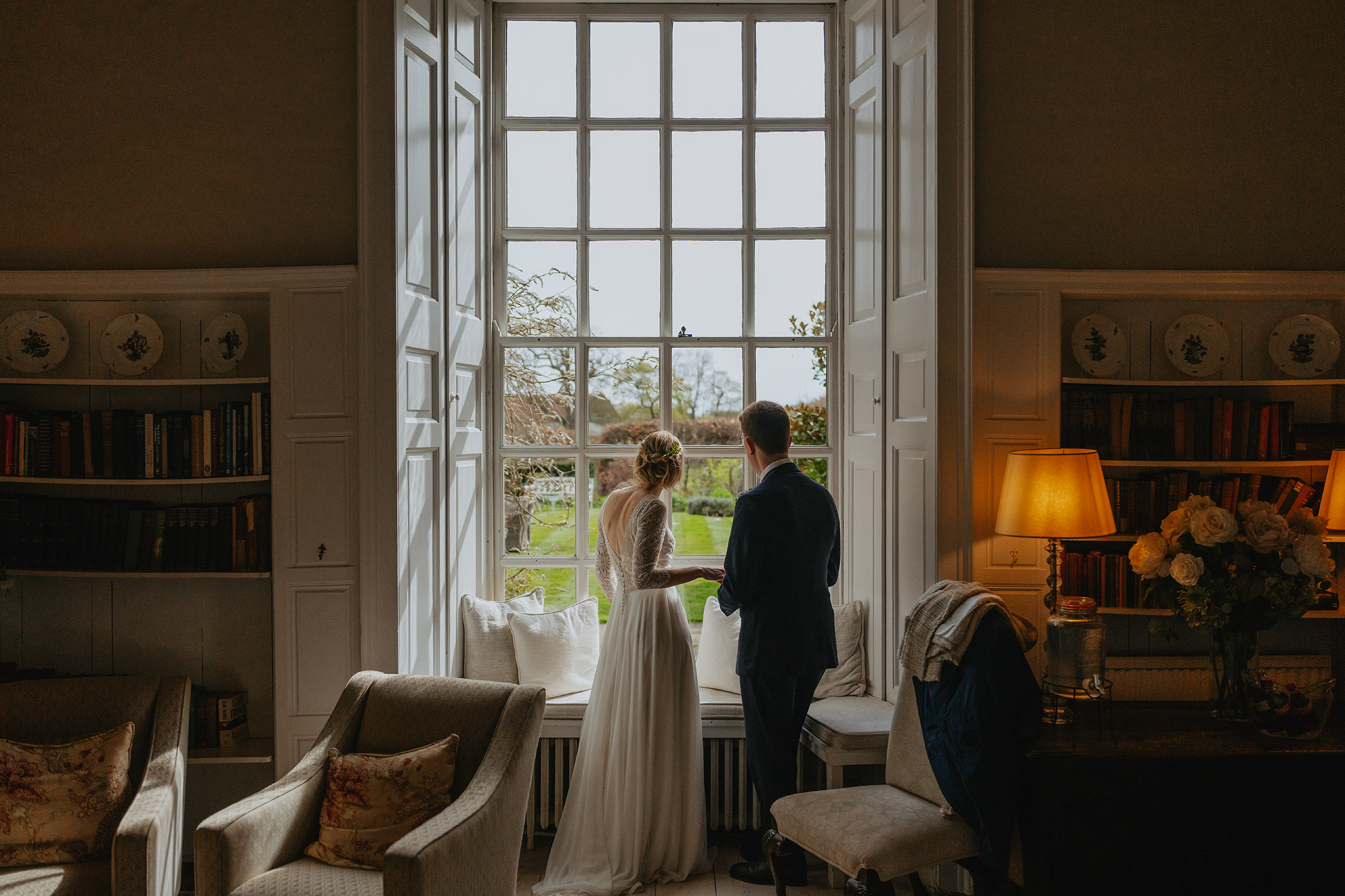 Couple in formal attire near a window, elegant room with shelves, viewing lush garden.