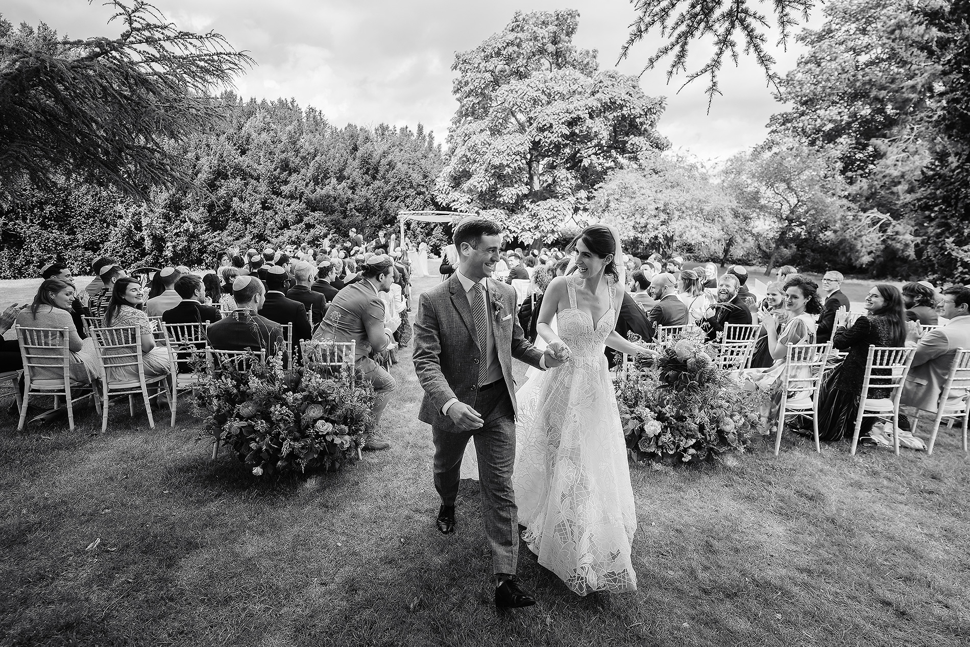 Couple holding hands in a garden ceremony with guests in formal attire.