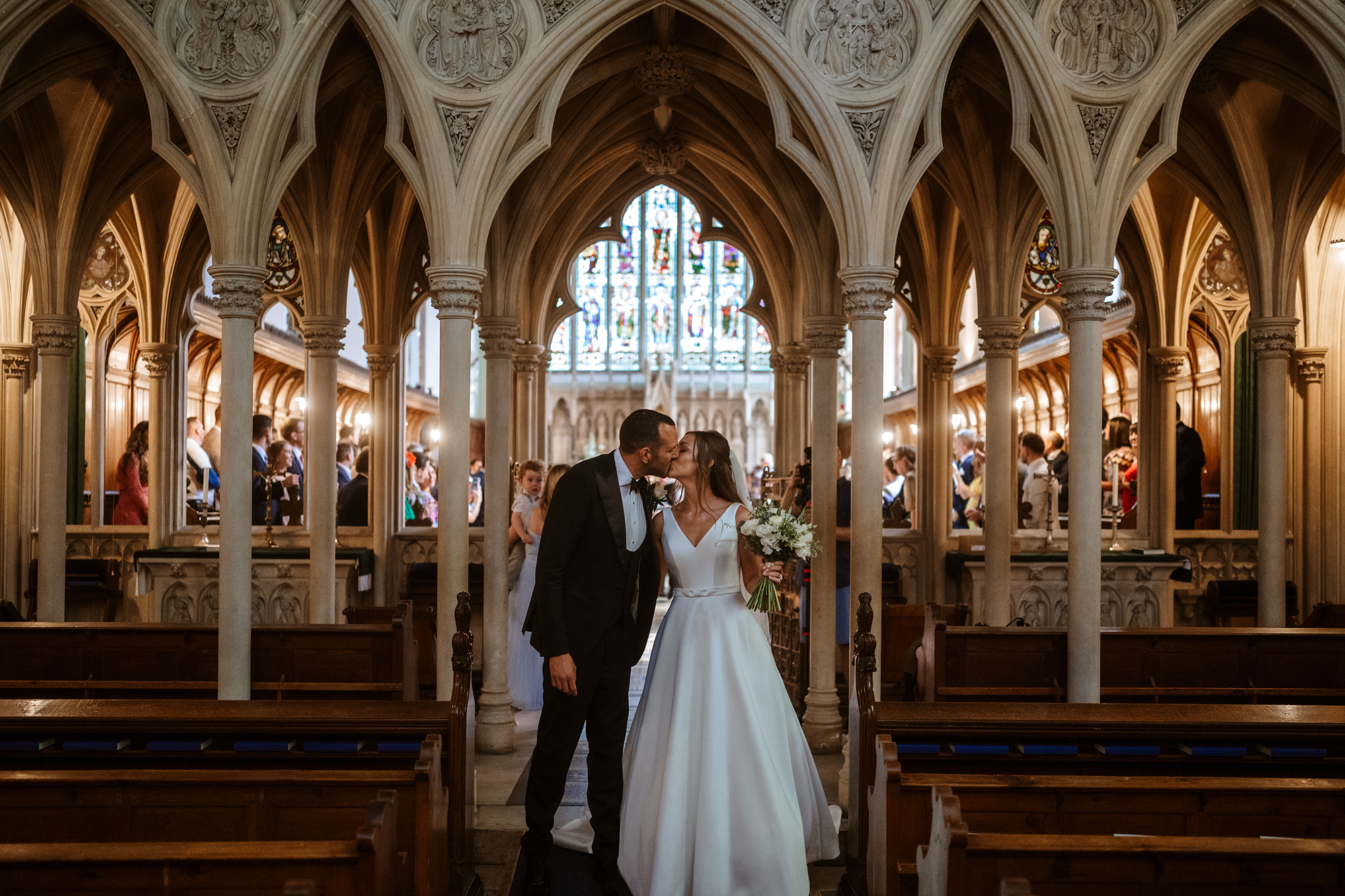 Couple kissing in church aisle under Gothic arches, bride in white dress, guests in background.