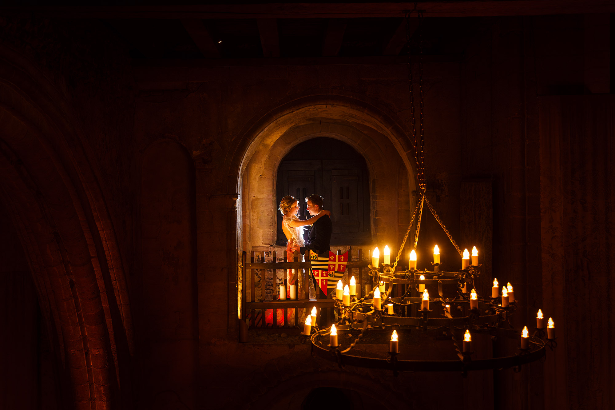 Couple embraces on a medieval balcony, lit by a glowing chandelier in a warm, dim scene.