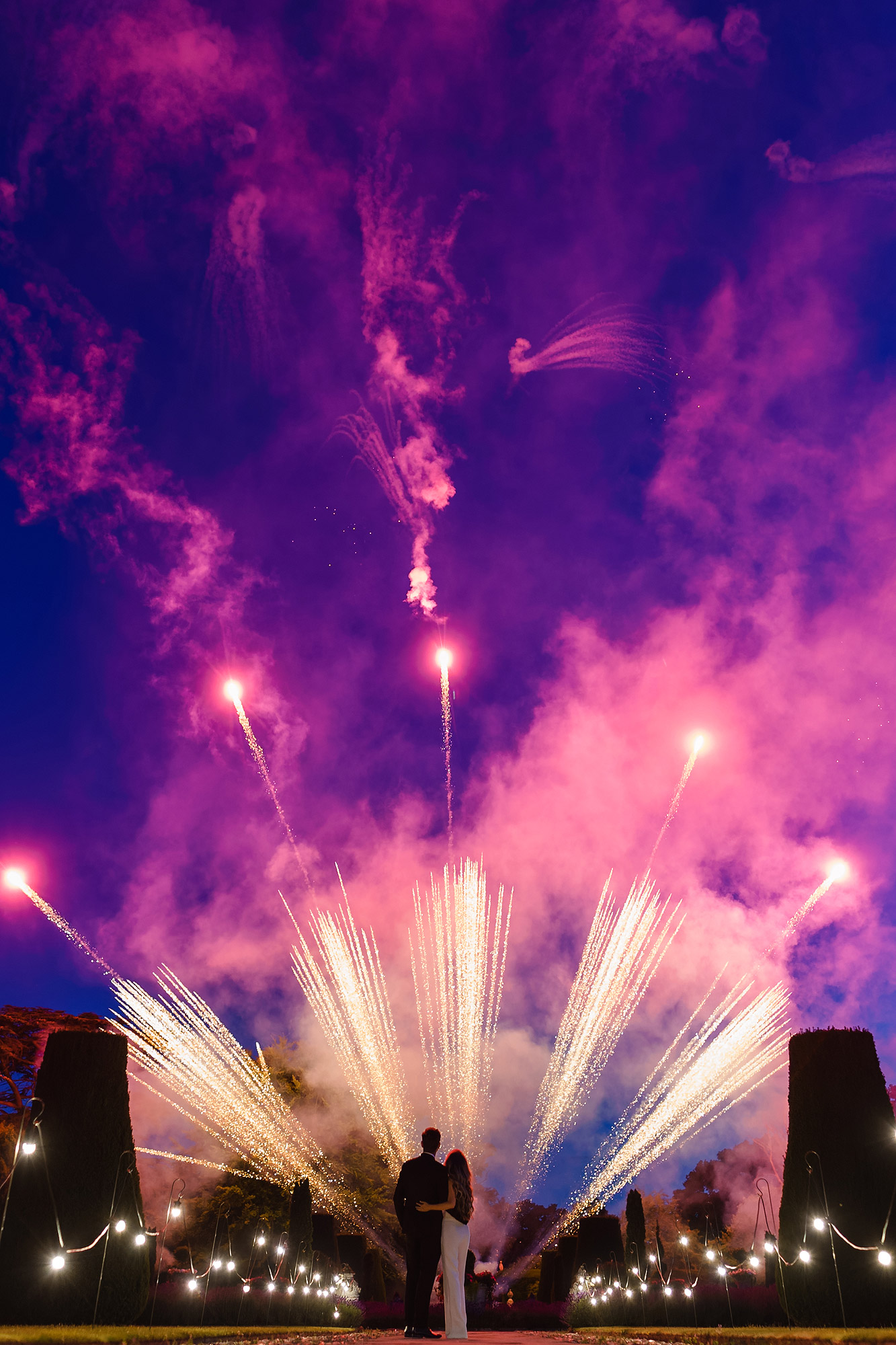Couple silhouetted by vibrant fireworks in a garden with decorative lights at night.