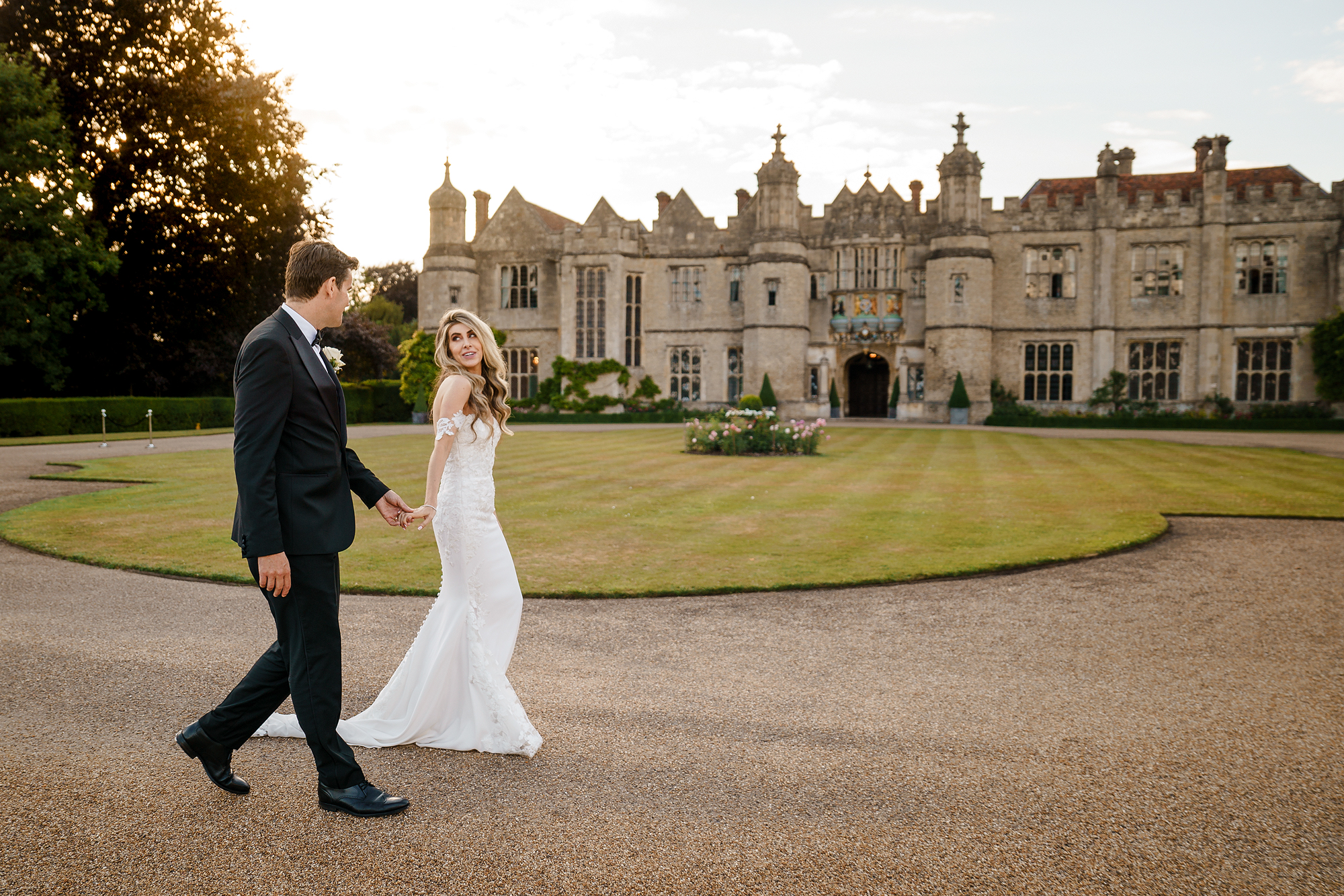 Couple in wedding attire walking on a path by a historic stone building with a lawn.