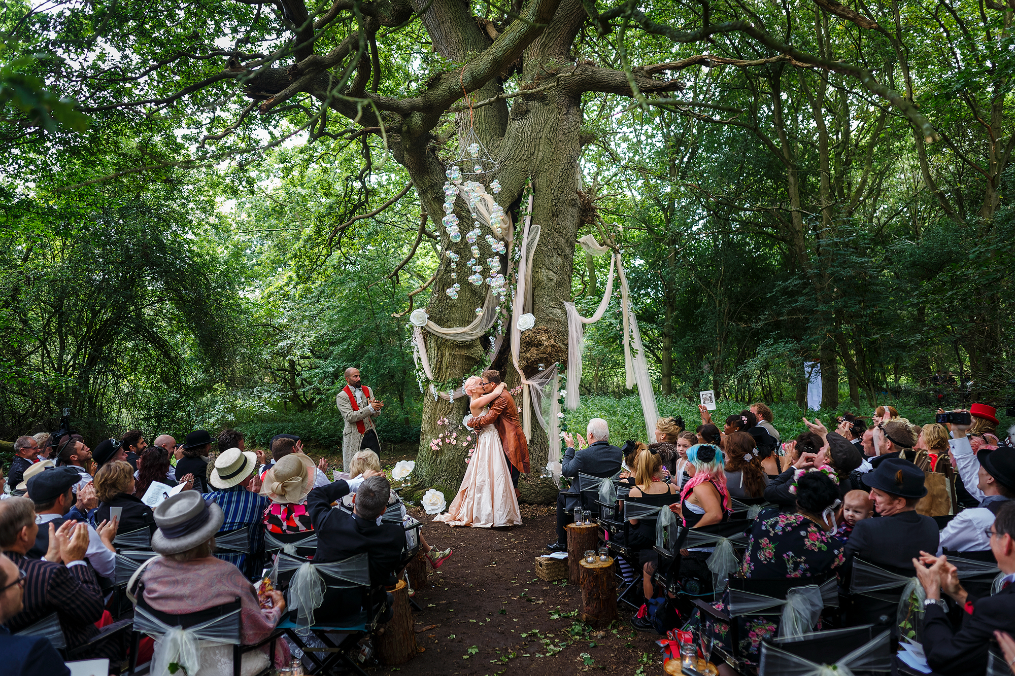 Wedding ceremony in forest with couple, guests, and decorations on large tree.