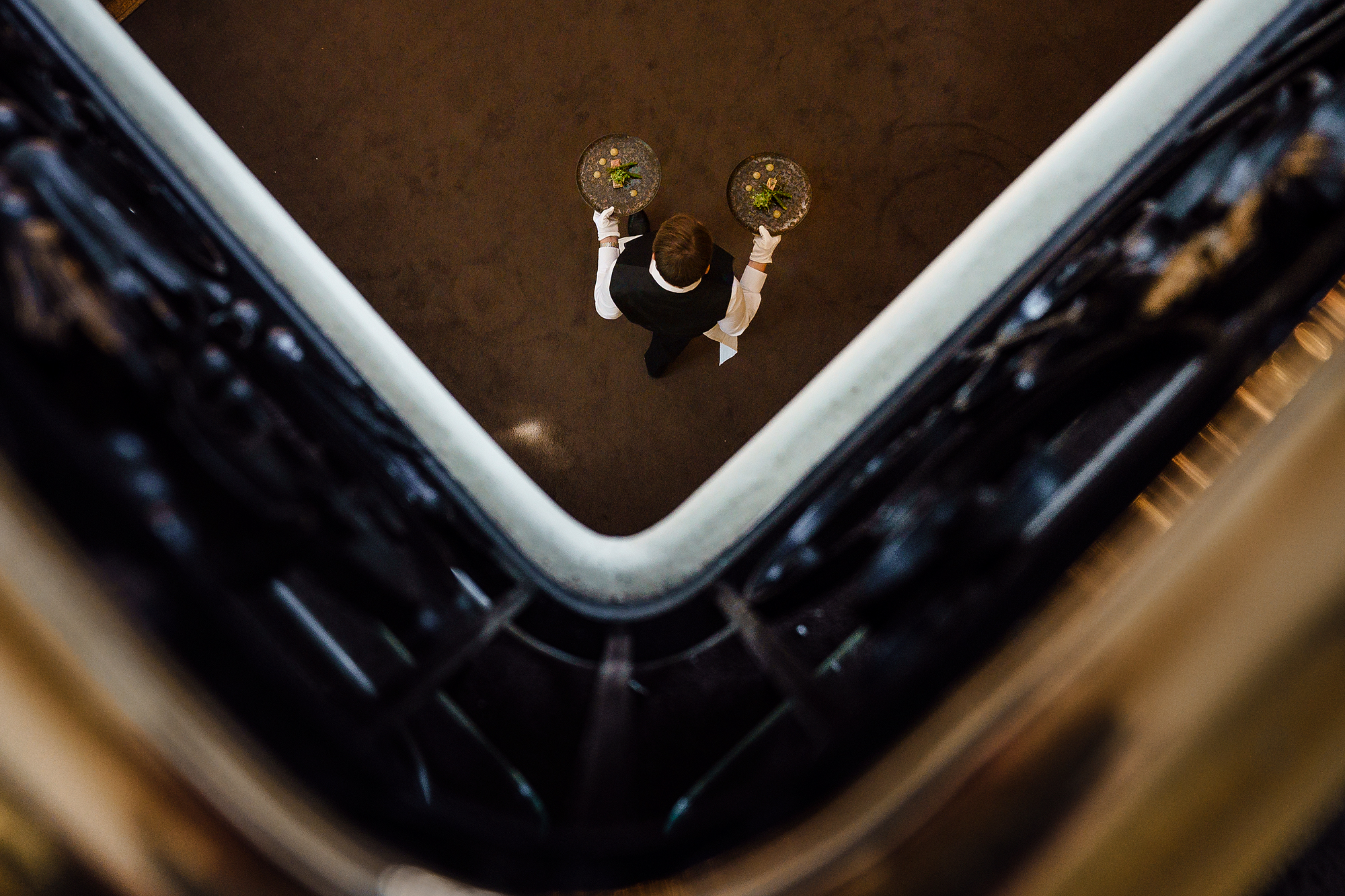 Person in formal attire carrying two food trays in elegant indoor space from above.