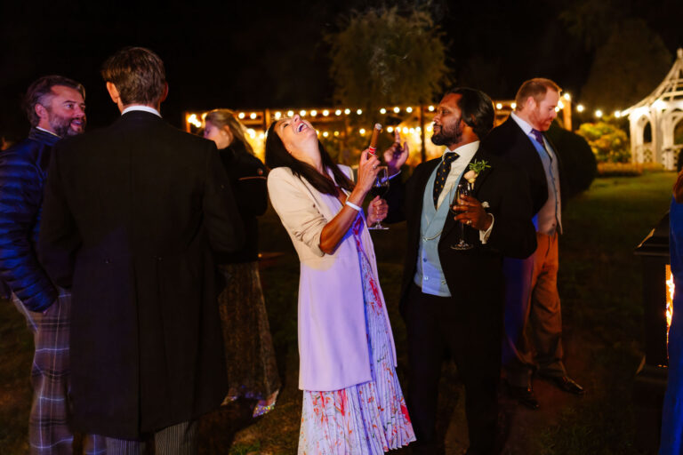 Group in formal attire laughing at outdoor night event with drinks and festive lights.