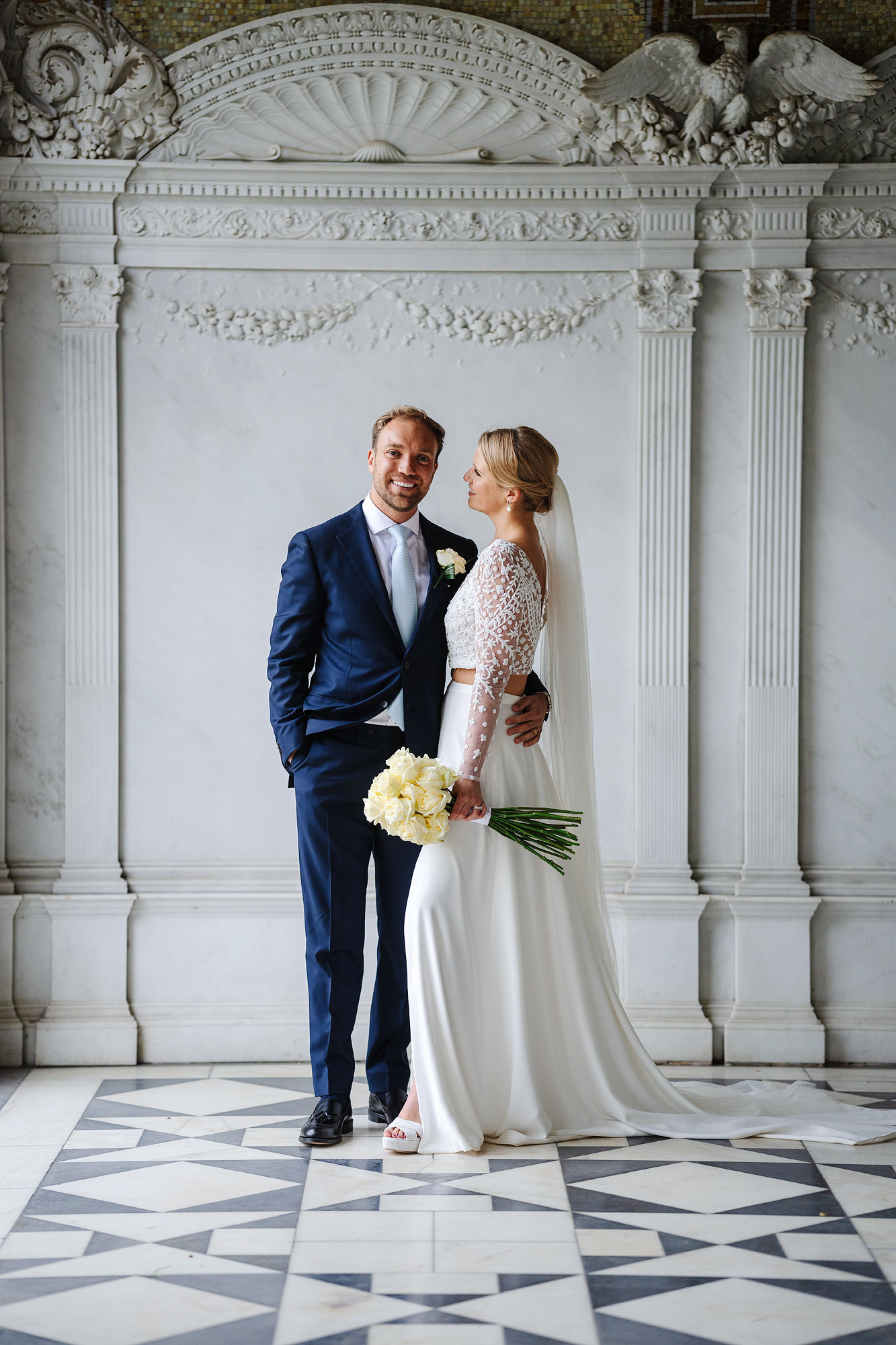 man in blue suit, woman in white gown with flowers, ornate backdrop.