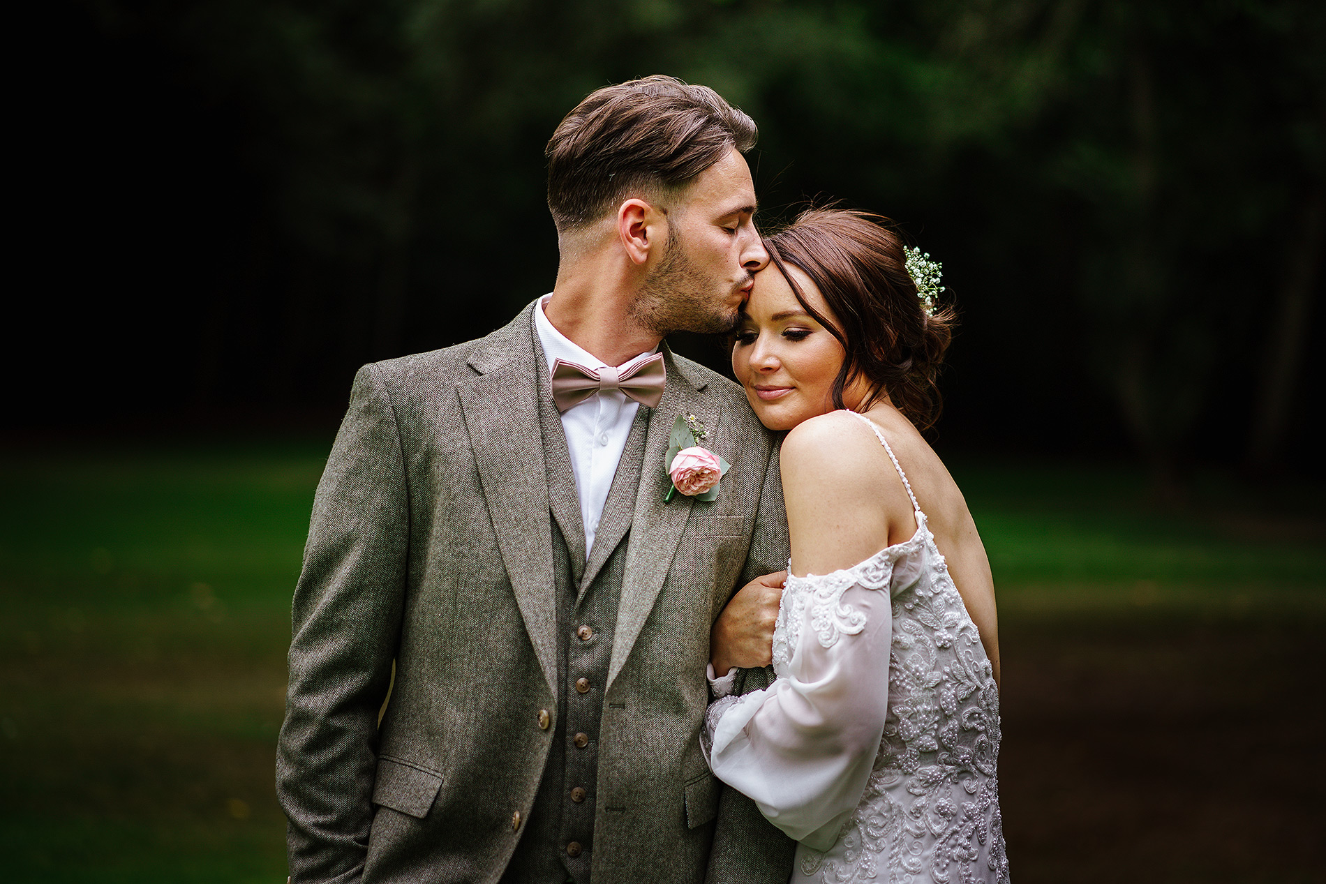 Man in grey suit kisses woman in white lace dress outdoors, resembling a wedding scene.