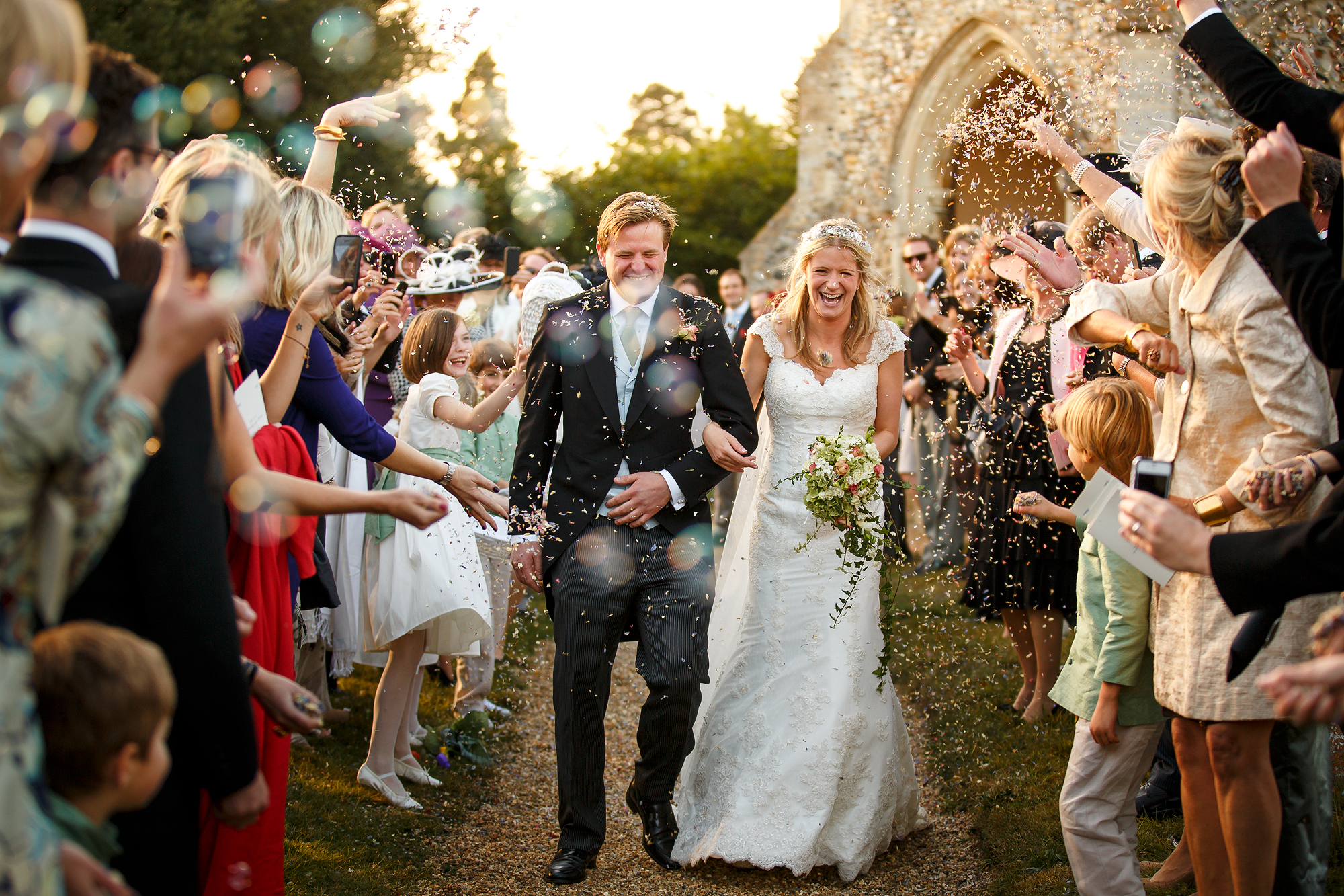 Newlywed couple walks through confetti, surrounded by smiling guests outdoors.