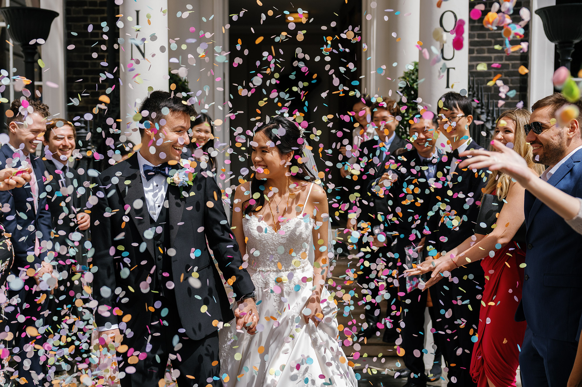 Newlywed couple in formal attire walking through colourful confetti with smiling guests.