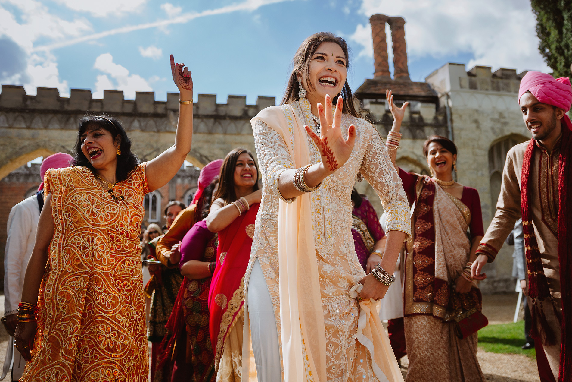 People dancing joyfully in colourful traditional attire, with henna and a historic building behind.