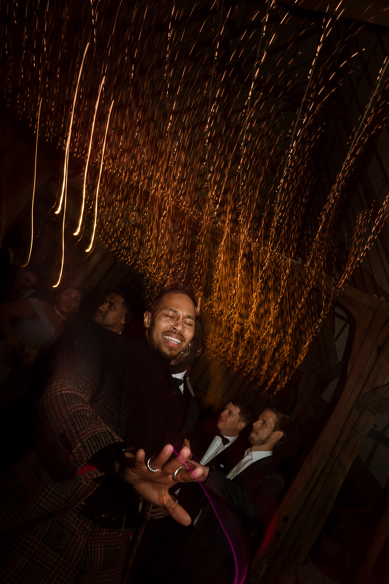 Person in suit and patterned coat dancing, blurred yellow lights above, others in background.
