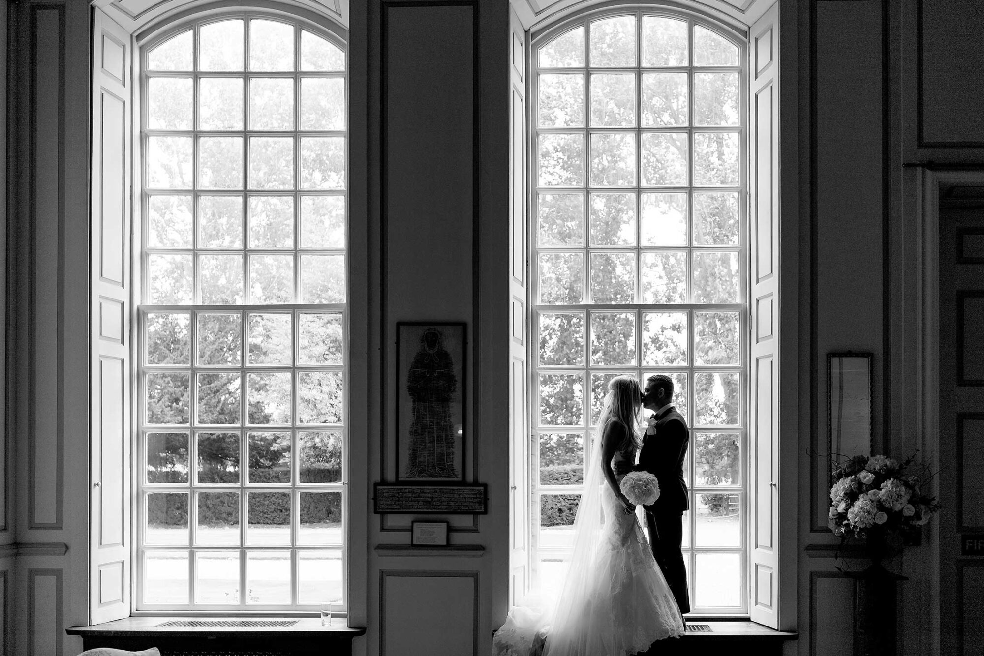 Silhouetted couple in wedding attire by large windows with trees visible outside.