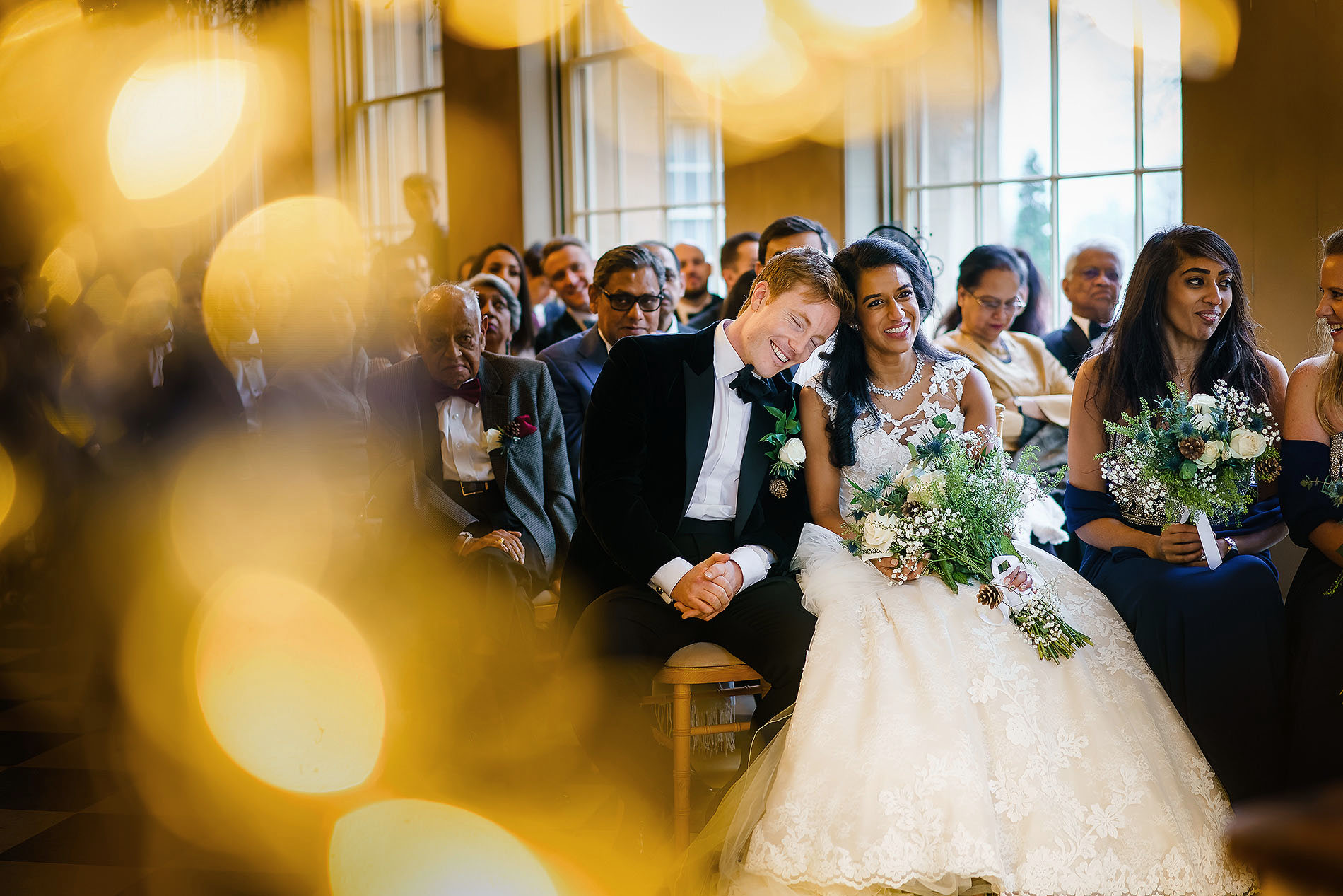 Wedding ceremony with a smiling bride and groom holding hands, surrounded by seated guests.