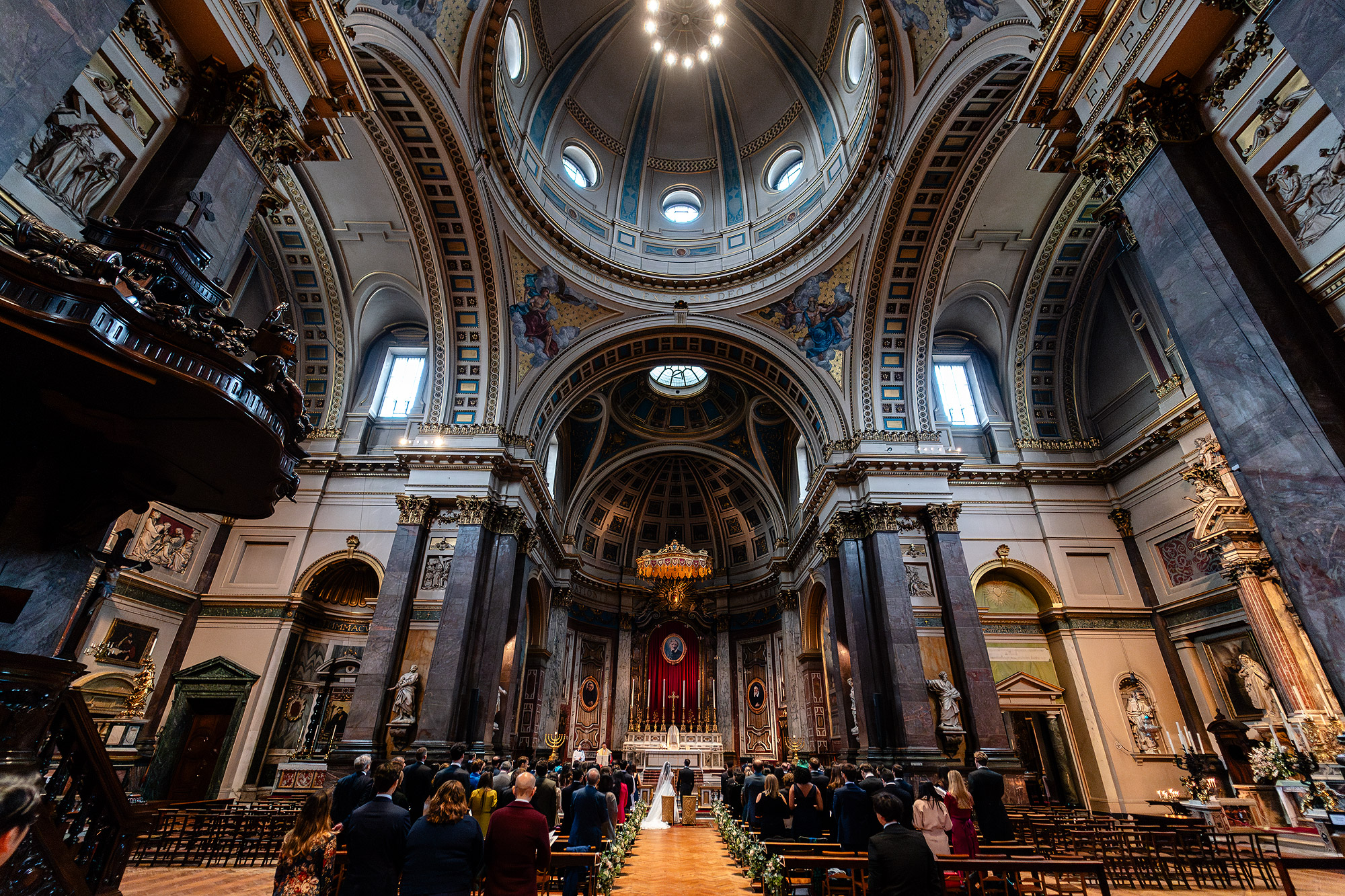 Wedding ceremony in an ornate cathedral with high dome, artwork, columns, and attendees.