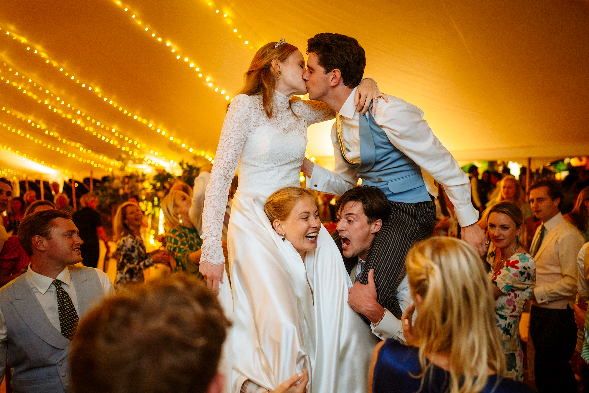 Wedding couple lifted on chairs kissing under tent with string lights.