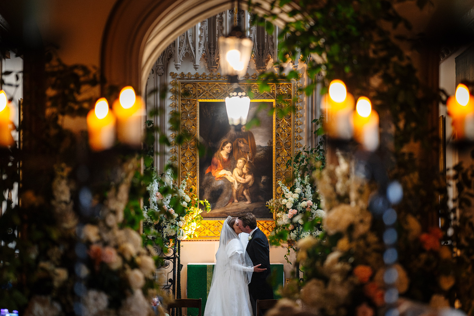 Couple in wedding attire kissing in a decorated church with flowers and candlelight.
