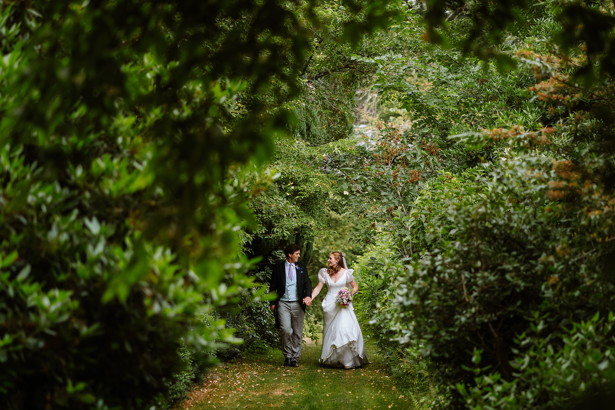 Couple in wedding attire walking through lush garden path, bride holding a flower bouquet.