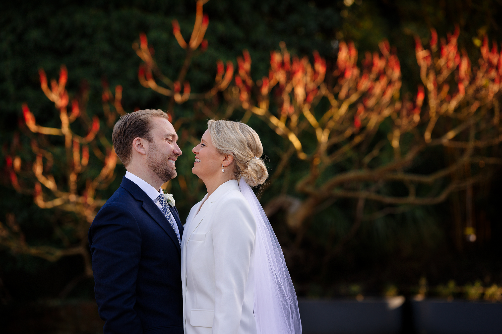 Couple smiles at each other outdoors, man in dark suit, woman in white with veil, greenery behind.