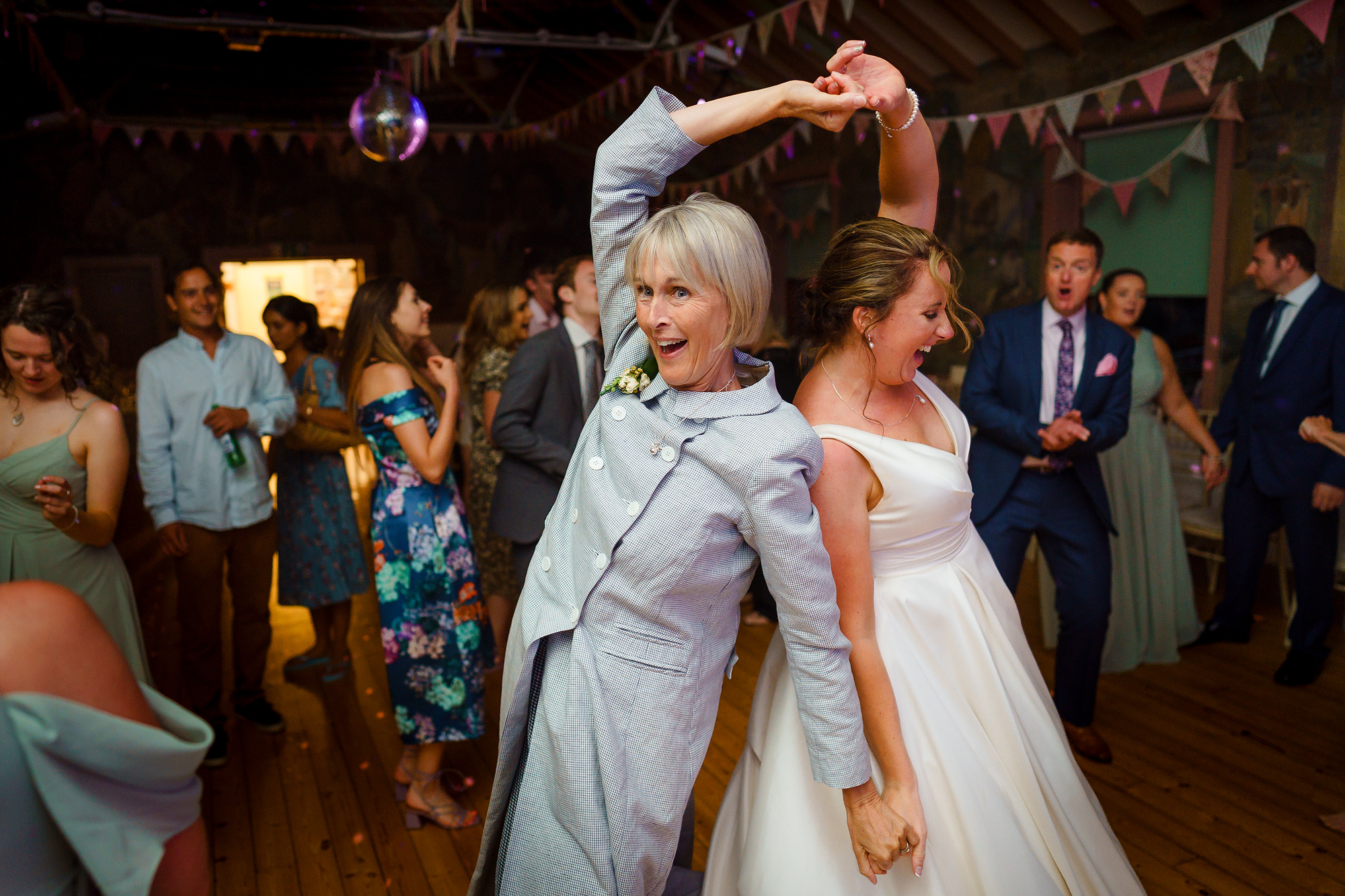 Wedding reception with guests dancing, a woman in grey and bride twirling, festive decor.