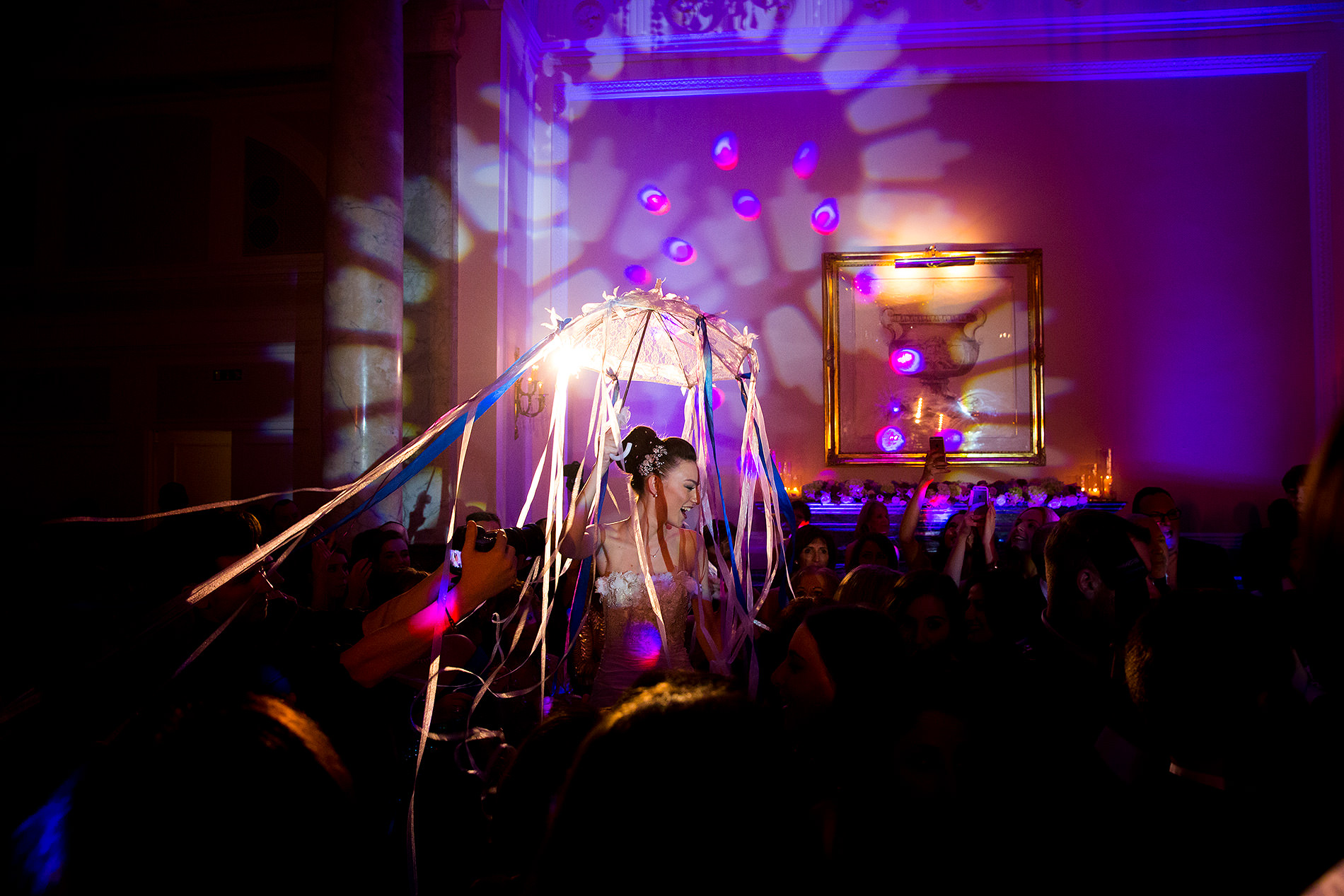Woman in formal attire under decorative canopy with ribbons, surrounded by crowd in festive room.